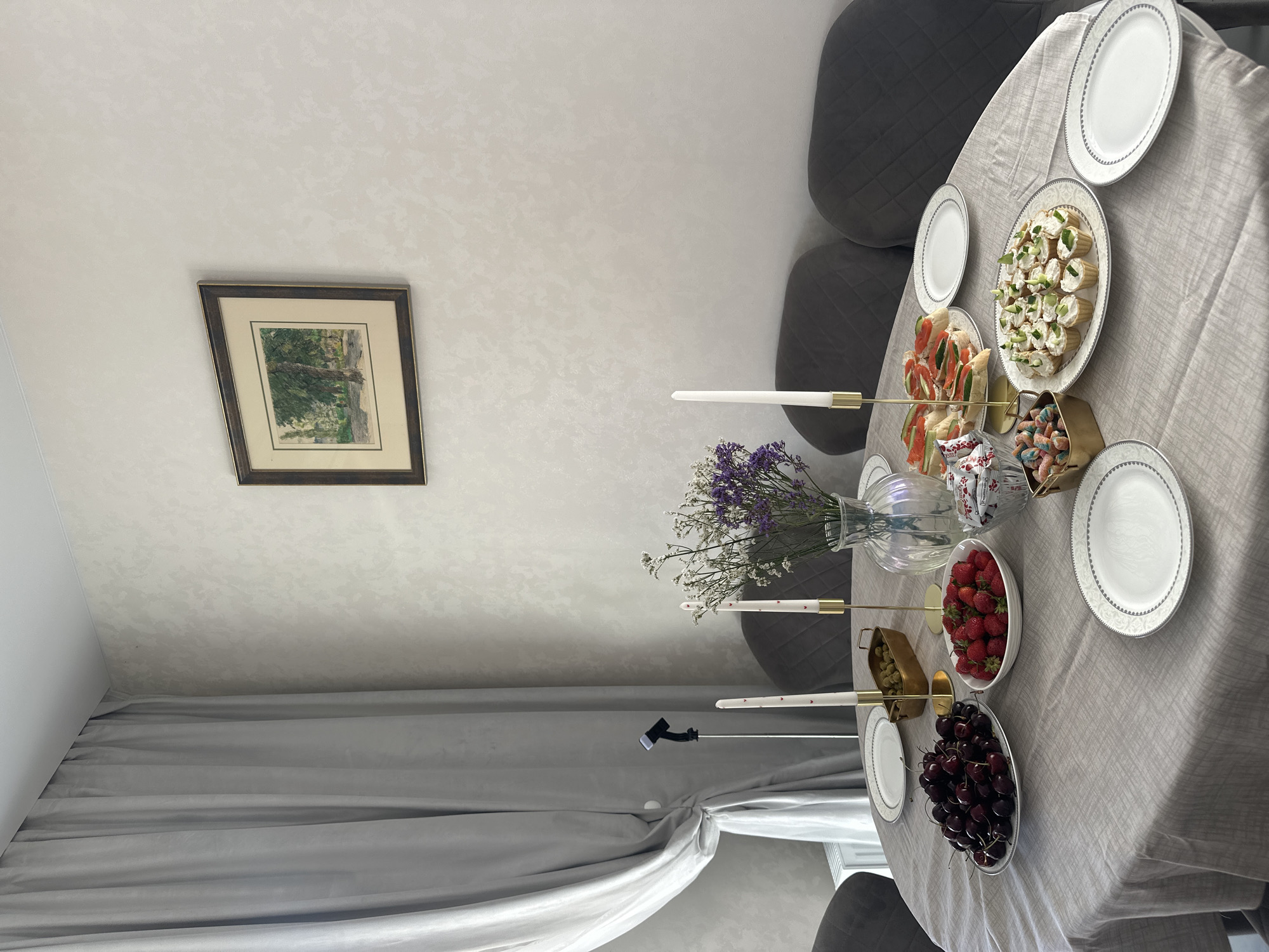 The image depicts a dining table set for a gathering, likely in a residential setting in Voronezh, Russia. The table is covered with a light-colored linen tablecloth. In the foreground, several white plates with a decorative rim are visible. To the left, a large platter is filled with dark cherries, and next to it, a bowl contains fresh strawberries. A golden rectangular dish holds green olives, and two tall white candles in golden holders stand in the center of the table. A glass vase with a bouquet of dried purple and white flowers adds a decorative touch. Further to the right, a plate of open-faced sandwiches with salmon and cucumber is arranged, accompanied by a small bowl of colorful candies. Another plate holds numerous small appetizer bites topped with cream cheese and cucumber. The background shows a textured light-colored wall with a framed picture of a park scene hanging on it. To the left, a grey curtain is drawn, and a black microphone stand with a microphone is positioned near the curtain. Dark grey upholstered chairs are visible around the table. The overall impression is a prepared meal with a variety of food items and decorative elements.