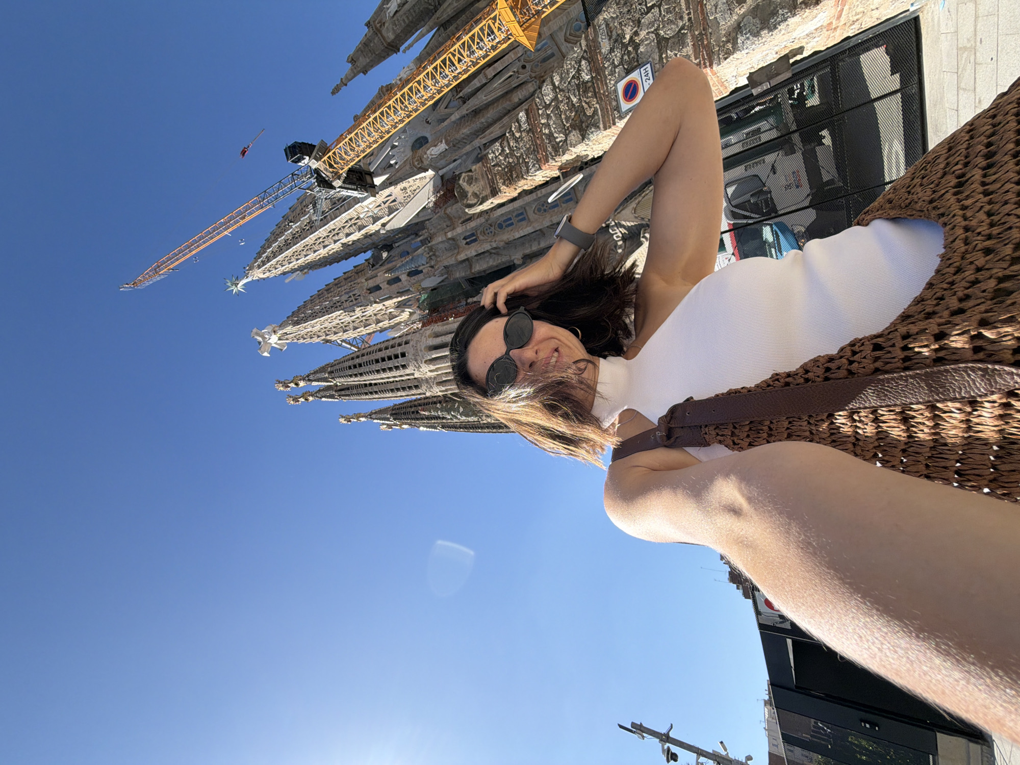 A woman is taking a selfie in front of the Sagrada Familia in Barcelona, Spain. She is wearing a white sleeveless top and sunglasses, and has a woven bag on her shoulder. The Sagrada Familia is a large, unfinished Roman Catholic minor basilica designed by Catalan architect Antoni Gaudí. Construction began in 1882 and is still ongoing. A large yellow crane is visible next to the basilica, indicating that construction is still in progress. The sky is clear and blue.