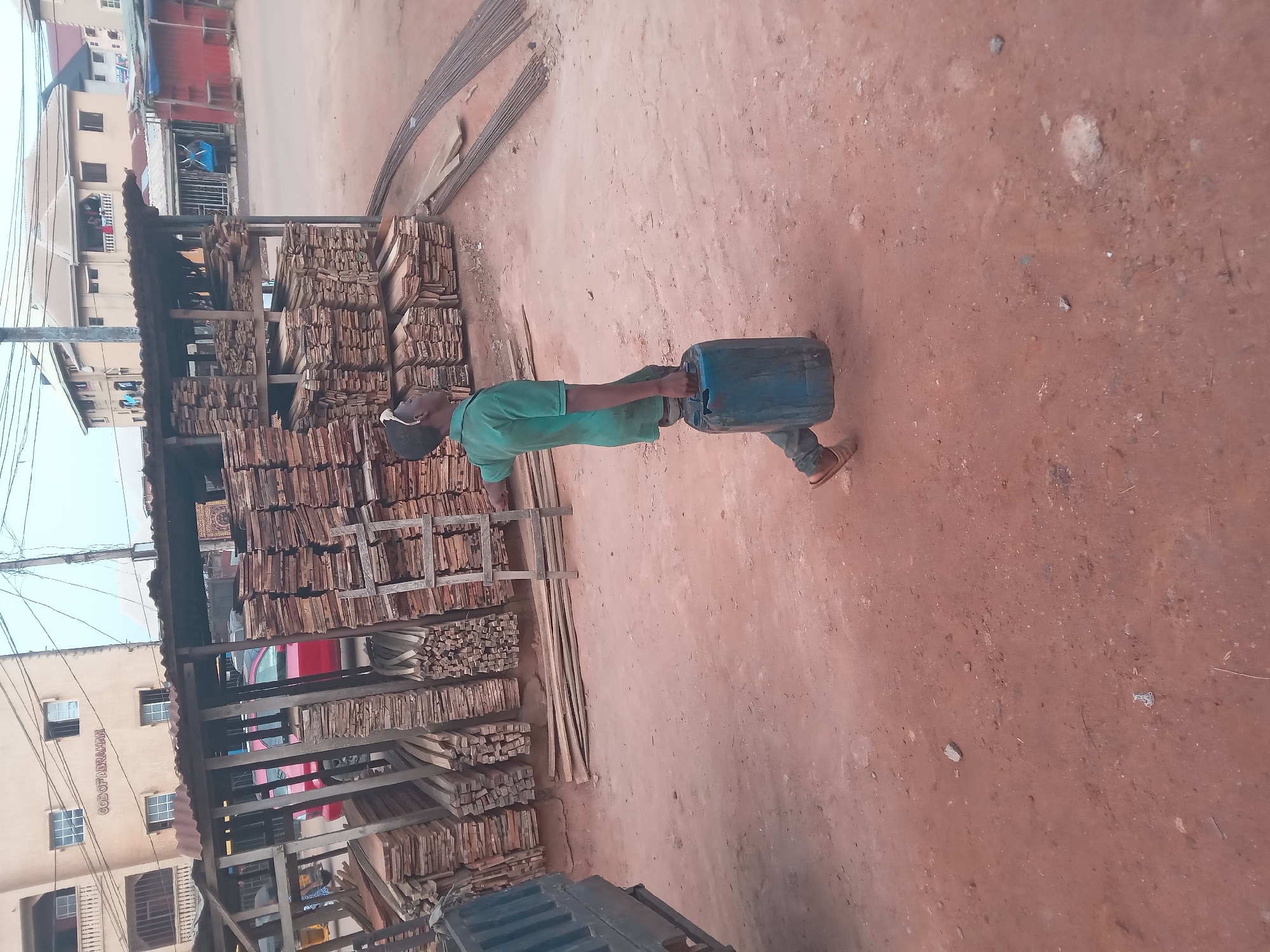 The image depicts a street in Nkpor, Nigeria, adjacent to a wood or lumber yard. A man in a green shirt carries a blue container. Stacks of lumber are visible under a corrugated metal roof. Rebar or metal rods are visible on the ground, and buildings are in the background. The ground is red dirt or soil.
