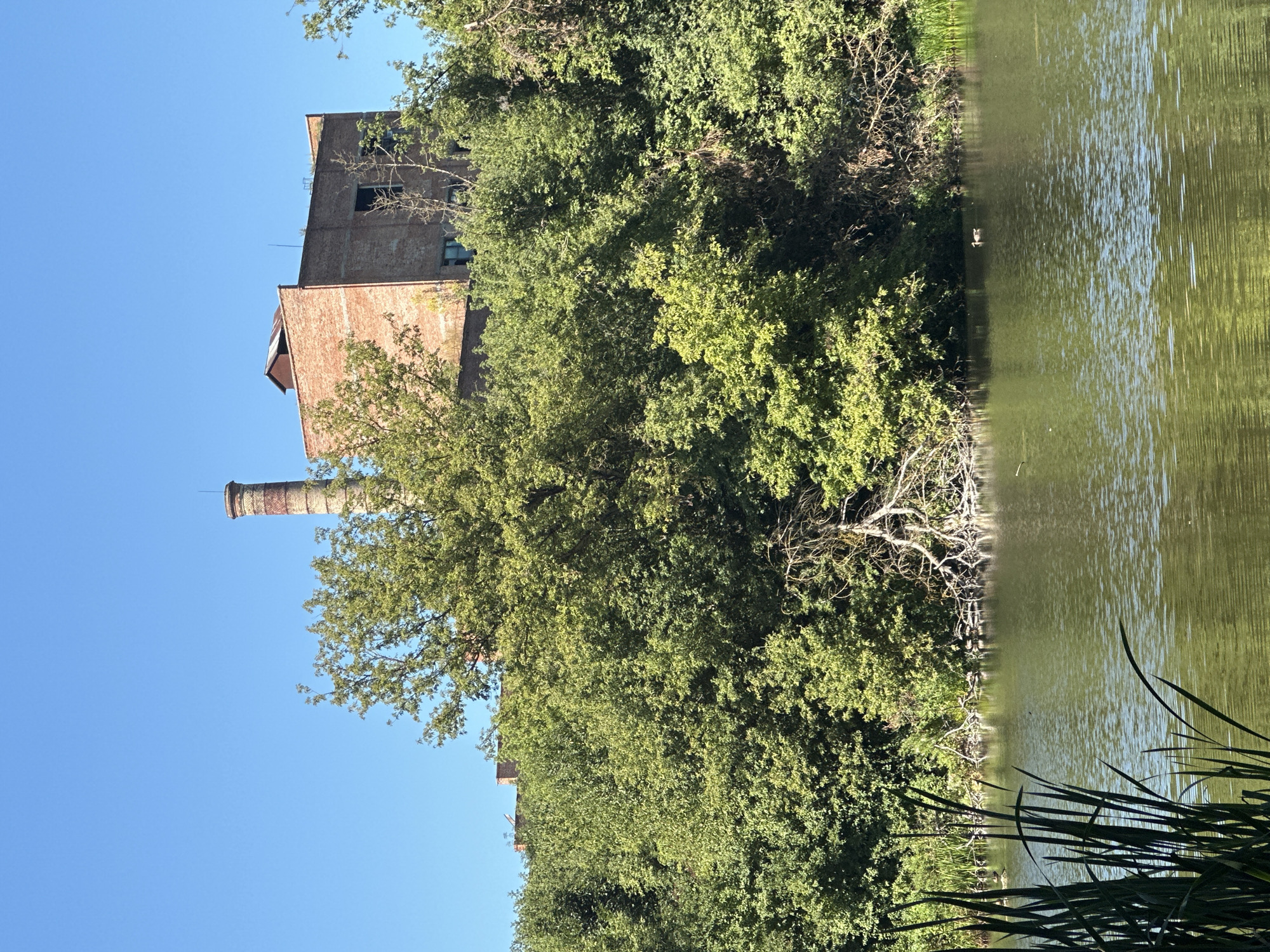 The image shows a brick industrial building with a tall smokestack, partially obscured by lush green trees, situated on a slight incline. In the foreground, a calm body of water reflects the trees and the blue sky. Several ducks are visible in the water. The scene appears to be in a semi-natural setting within or near the city of Makiv, Ukraine.