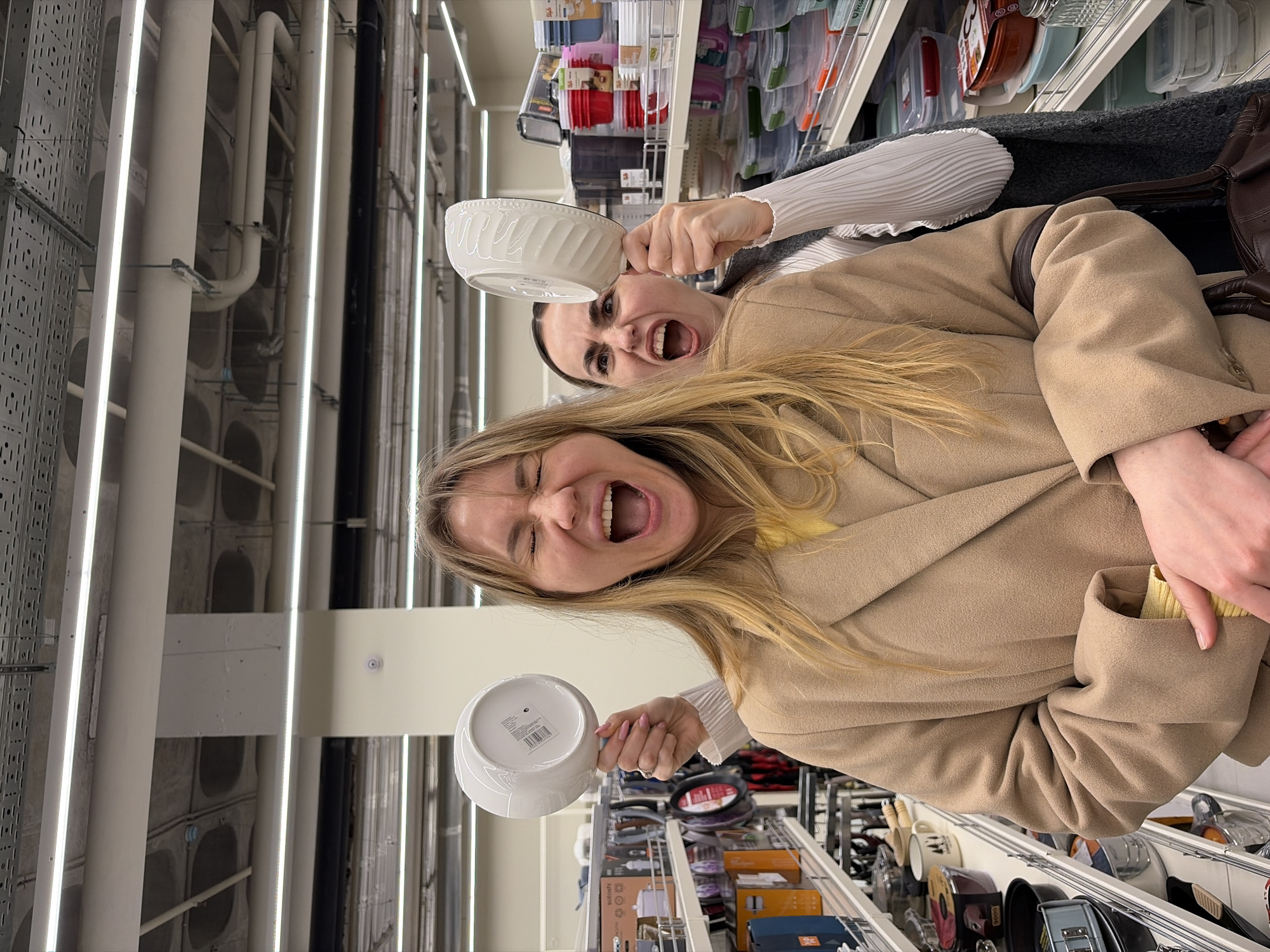 Two women are standing in a store aisle, possibly in the city of Izhevsk, Russia. The woman in the foreground has long blonde hair and is wearing a tan coat and yellow sweater. She is holding a white bowl and yelling. The woman behind her has dark hair and is wearing a gray sweater with a white long-sleeved shirt underneath. She is also holding a white bowl and yelling. The aisle is lined with shelves of various kitchen items and storage containers. Overhead are white pipes and fluorescent lights.
