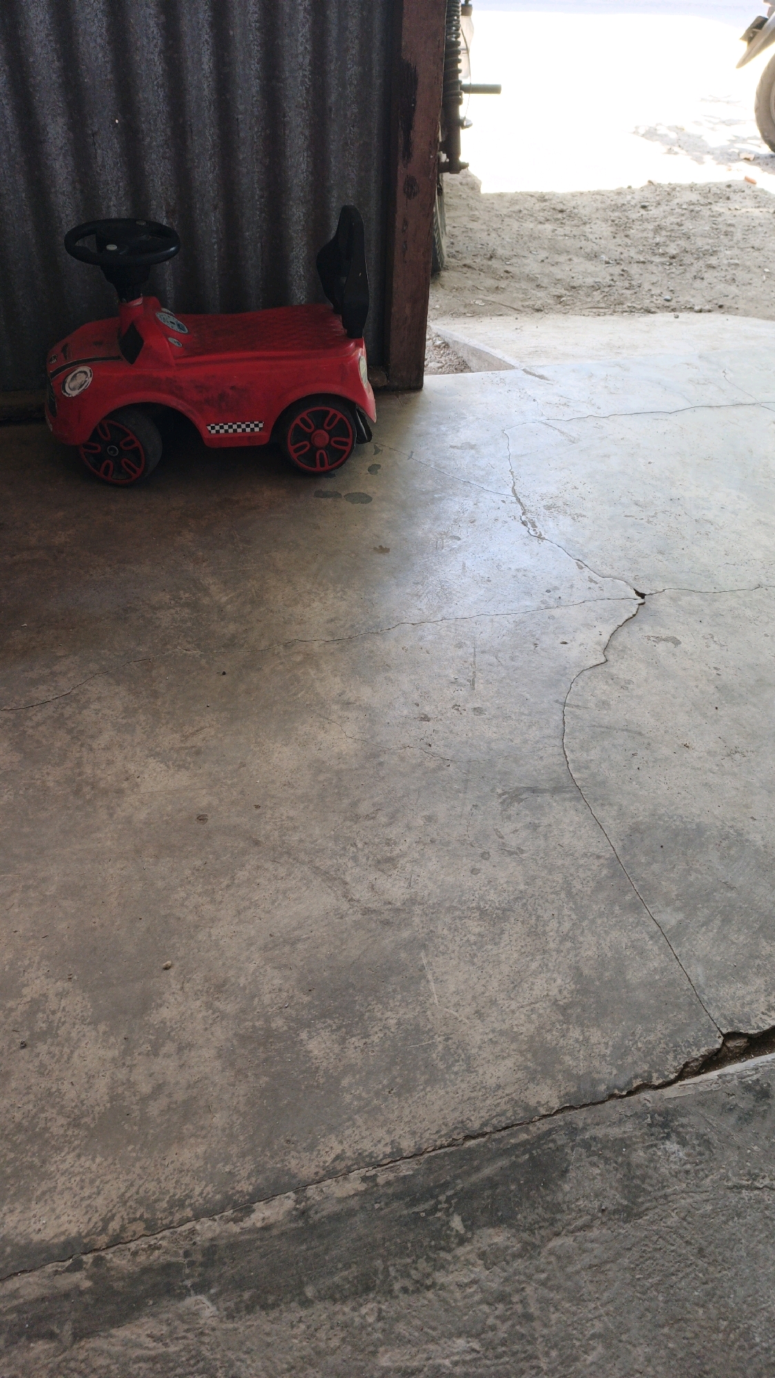 A red toy car with black wheels and a black steering wheel sits on a concrete floor. The car has a checkered pattern on its side and what appears to be a logo on the hood. To the left of the car is a corrugated metal wall. To the right of the car, the concrete floor shows significant cracks. The background is a bright, outdoor area with dirt and debris, suggesting an open space or possibly a construction site. This scene appears to be located outdoors in the city of Kiping, Indonesia.