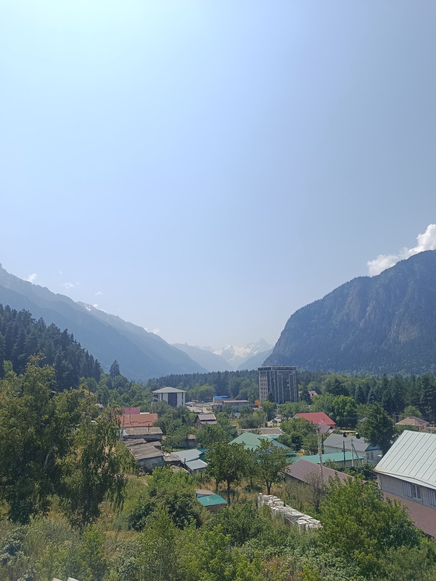 The image depicts a wide shot of the city of Teberda, Russia, nestled within a valley surrounded by majestic mountains. The sky is a clear, pale blue, with a few wispy clouds visible towards the right. In the foreground, a lush green landscape dominates, with dense trees and bushes covering the terrain. Various houses, with roofs of different colors and materials, are scattered throughout the scene, indicating a residential area. Some of the houses appear to be older, with corrugated metal roofs, while others have more modern designs. A tall, multi-story building stands out among the houses, suggesting a more urbanized section of the city. The background is characterized by the imposing presence of mountains, with forested slopes leading up to rocky peaks. Some of the distant mountains show hints of snow, even under the bright daylight. The overall impression is of a peaceful town integrated with its natural mountainous environment.