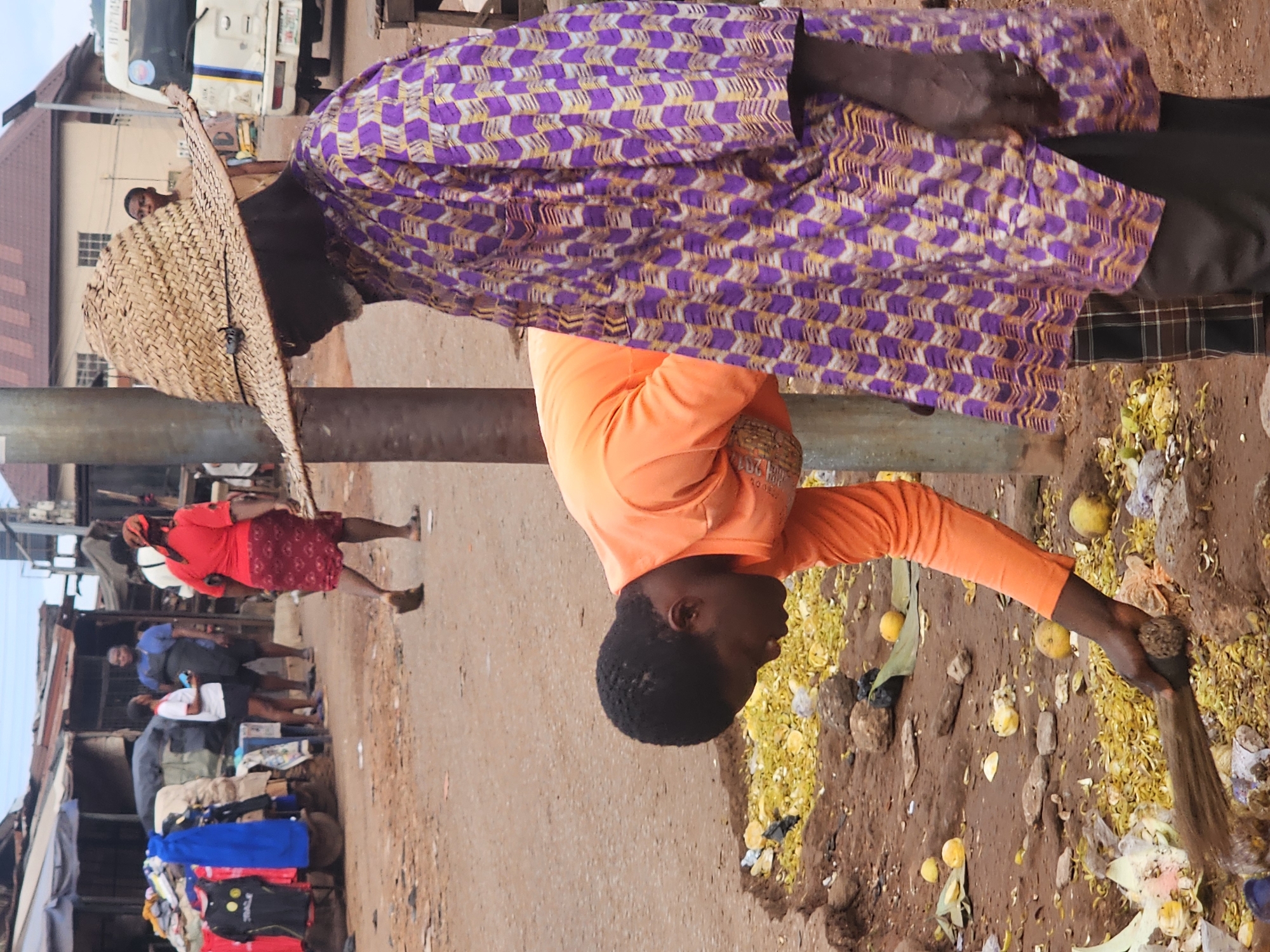 A low-angle, medium shot captures a street scene with two individuals in the foreground and a bustling market in the background. The ground is covered in dirt, debris, and scattered yellow fruit, suggesting an outdoor setting.
In the immediate foreground, to the left, a person wearing an orange long-sleeved shirt is bending down, sweeping the ground with a broom. Their body is angled away from the camera, with their head lowered.
To the right of the sweeping individual, a second person is partially visible, wearing a straw hat and a vibrant purple and yellow patterned garment. They appear to be standing still.
In the background, several stalls with rustic roofs are set up, displaying various items including clothing. People are seen walking around and interacting within the market. A white vehicle with blue and yellow stripes is parked on the right side of the frame. The sky above is overcast.