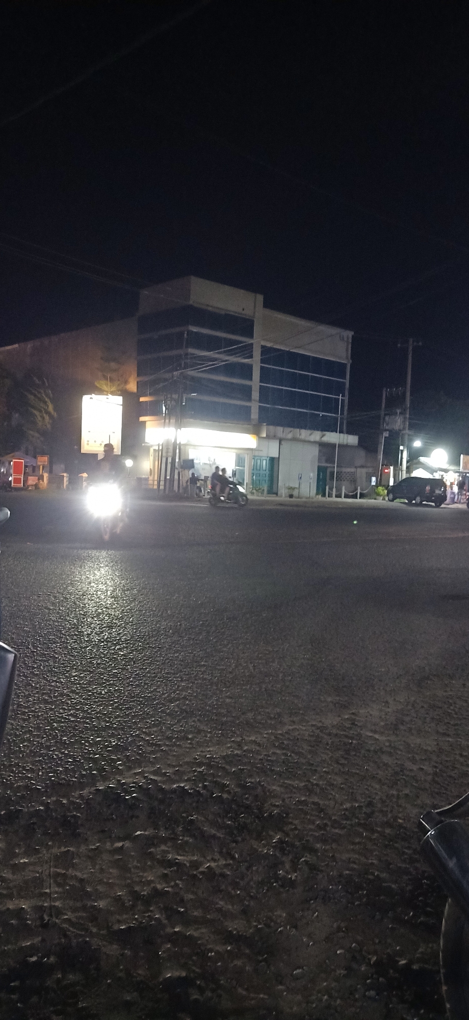 A wide shot captures a street scene at night in Lhokseumawe, Indonesia, likely near the intersection of Jalan Merdeka and Jalan Listrik. The foreground is dominated by a dark, wet asphalt road, reflecting the ambient light from streetlights and passing vehicles. In the midground, a modern, multi-story building with a reflective glass facade stands prominently. Its lower level appears to be commercial, with bright lights illuminating storefronts. To the left of the building, a large illuminated billboard is visible.
Several people are present, primarily on the road and sidewalks. A prominent subject is a motorcycle with its headlight on, approaching the camera from the left, with its rider silhouetted against the bright light. Further into the scene, another motorcycle carries two passengers, moving away from the camera towards the building. A dark-colored car is parked or moving on the right side of the road. The environment is characteristic of a developing urban area, with utility poles and wires crisscrossing the sky, indicating the presence of infrastructure. The overall impression is a bustling intersection at night, with the architecture suggesting a commercial or administrative center within the city.