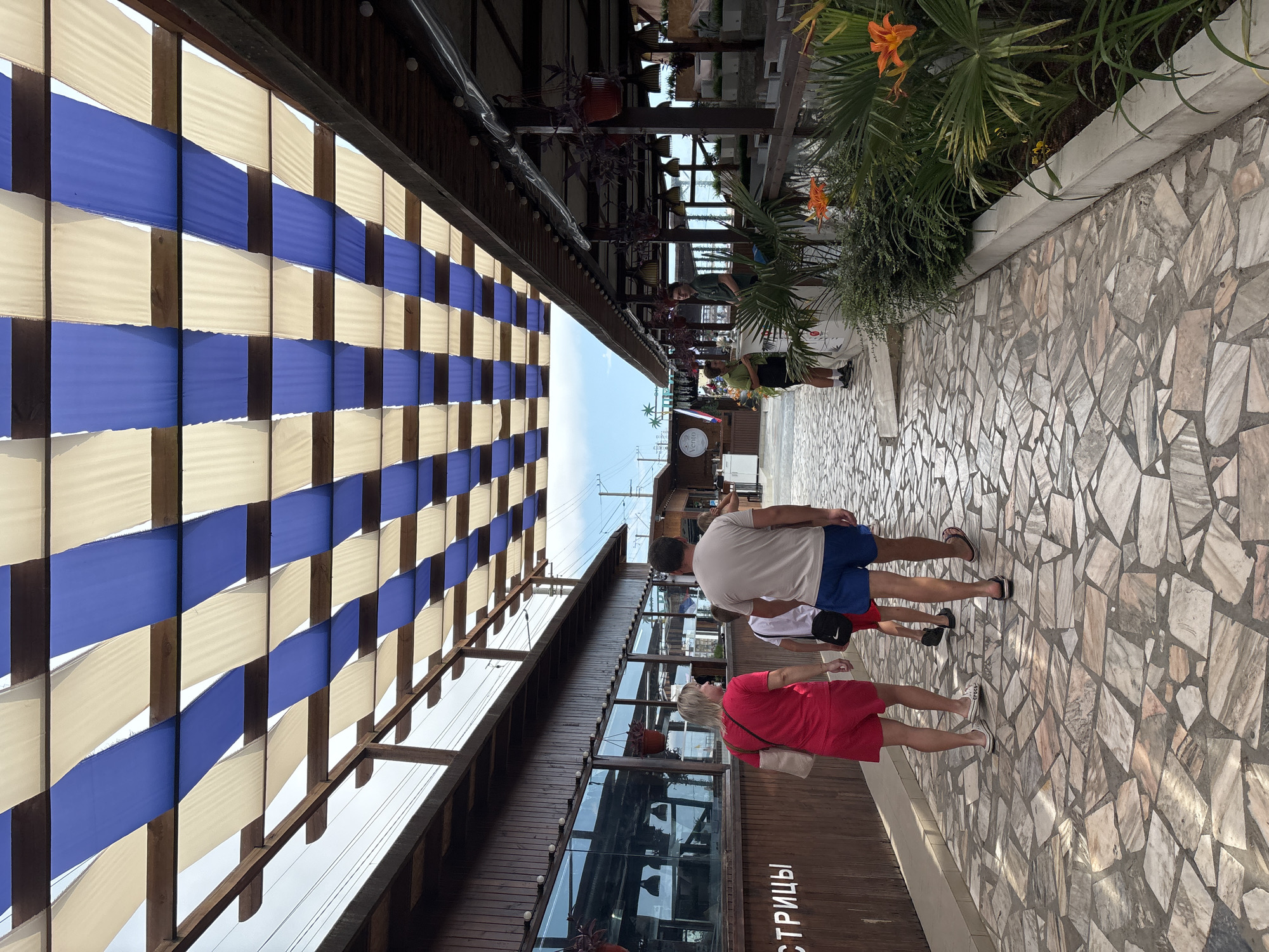 A family of four is walking down a paved walkway lined with buildings and an outdoor seating area. The walkway is made of irregularly shaped stone tiles with a textured surface. The buildings on either side are rustic, with wooden facades. The building on the left has large windows, and the text "СТРИЦЫ" is visible on its wall. The outdoor seating area on the right is covered by a canopy made of alternating blue and white fabric strips stretched over a wooden frame. The canopy provides shade to the seating area, which has tables and chairs. In the background, palm trees and the sky are visible, suggesting a warm climate. The scene appears to be a promenade or outdoor dining area, likely in a resort town. The clothing of the people, such as shorts and light shirts, indicates warm weather. A Russian flag is visible on a building in the distance on the right side of the image, potentially indicating the location is within Russia. The overall atmosphere is casual and relaxed.