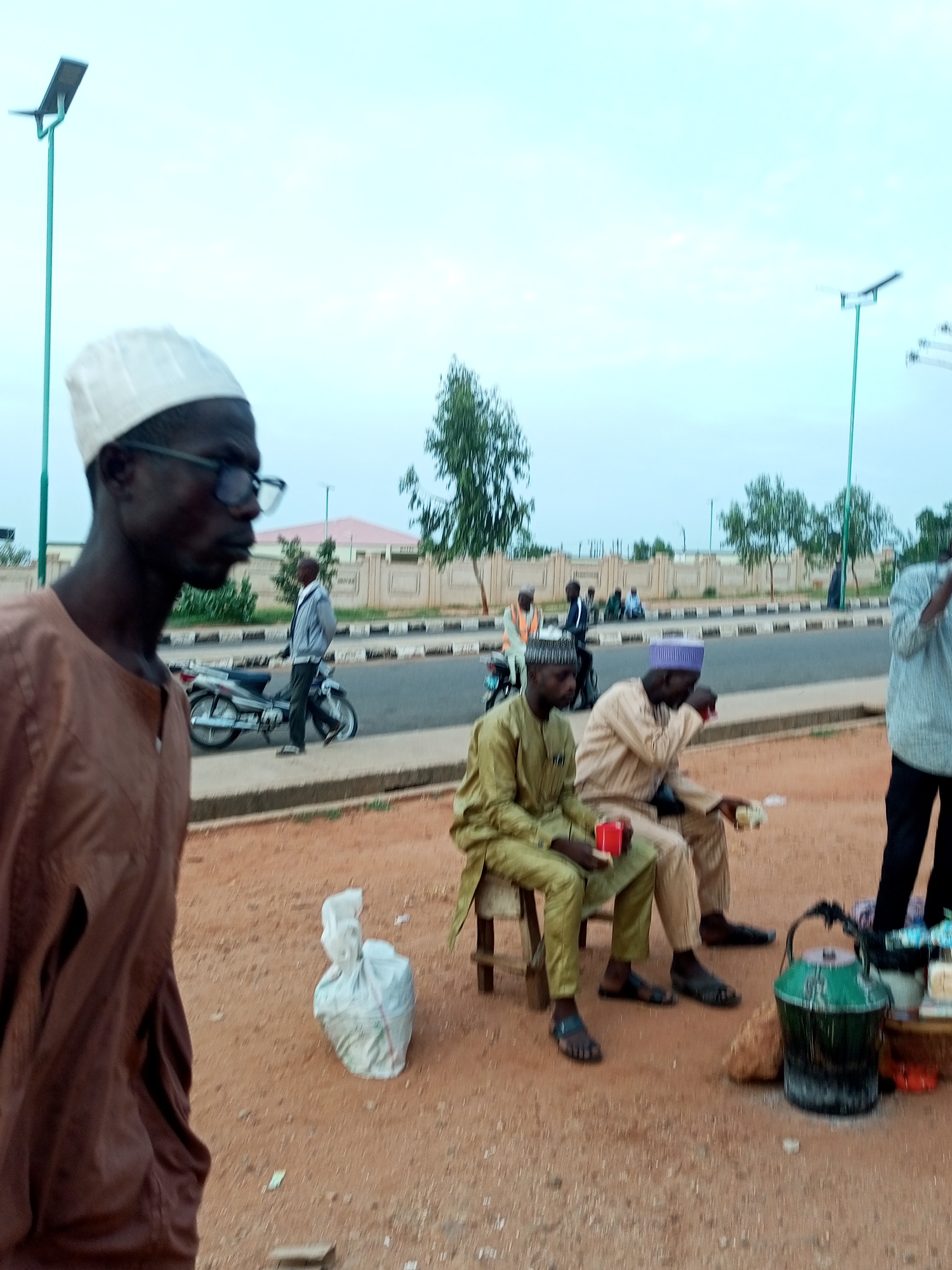 A street scene in Gombe, Nigeria, depicts individuals gathered near a road. In the foreground, a man wearing a brown robe and a white cap stands looking to the left. Further back, two men are seated on chairs by the roadside. One, wearing a light green kaftan and a cap, holds a red cup. The other, in a tan kaftan and a purple cap, is also holding a cup and appears to be eating. A green bucket and other items are arranged near them. A motorcycle is parked on the side of the road, with a man in a light jacket standing next to it. In the background, a building with a pink roof is visible, along with trees and streetlights. The ground is a reddish-brown dirt.