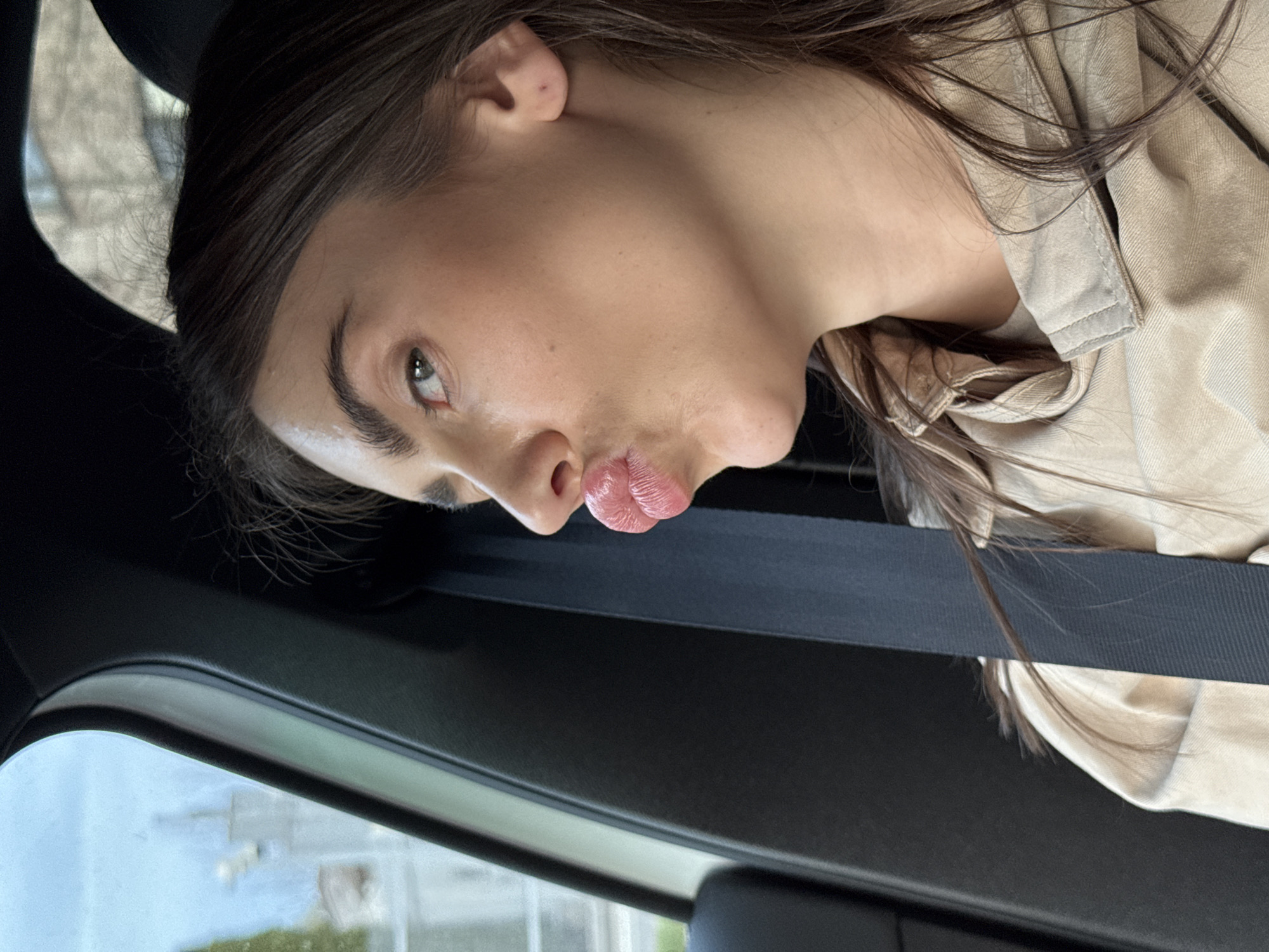 A young adult female is sitting in a car, wearing a tan jacket and a seatbelt. The vehicle is possibly in Warsaw, Poland, inferred from the blurred cityscape and architecture visible through the car window. The woman is looking towards the camera with pursed lips.