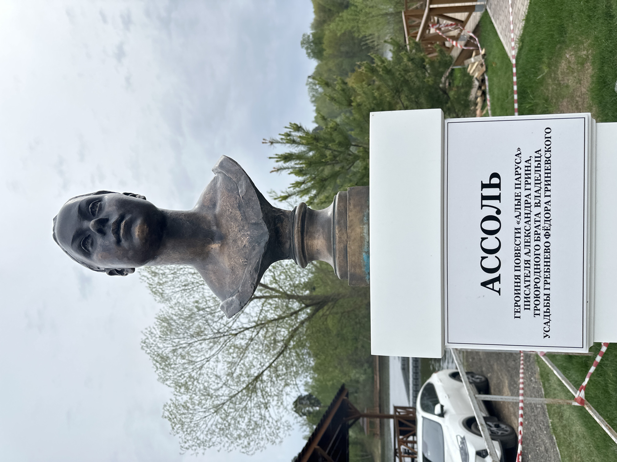 The image shows a bronze bust of Assol, the main character in Alexander Grin's novel "Scarlet Sails," located in Grebnevo, Russia. The bust is mounted on a pillar, next to a sign that identifies Assol as the heroine of the novel and mentions the connection between the author and the owner of the Grebnevo estate. The surroundings include trees, grass, a wooden structure, a car, and a red-and-white striped barrier tape. The sky is overcast.