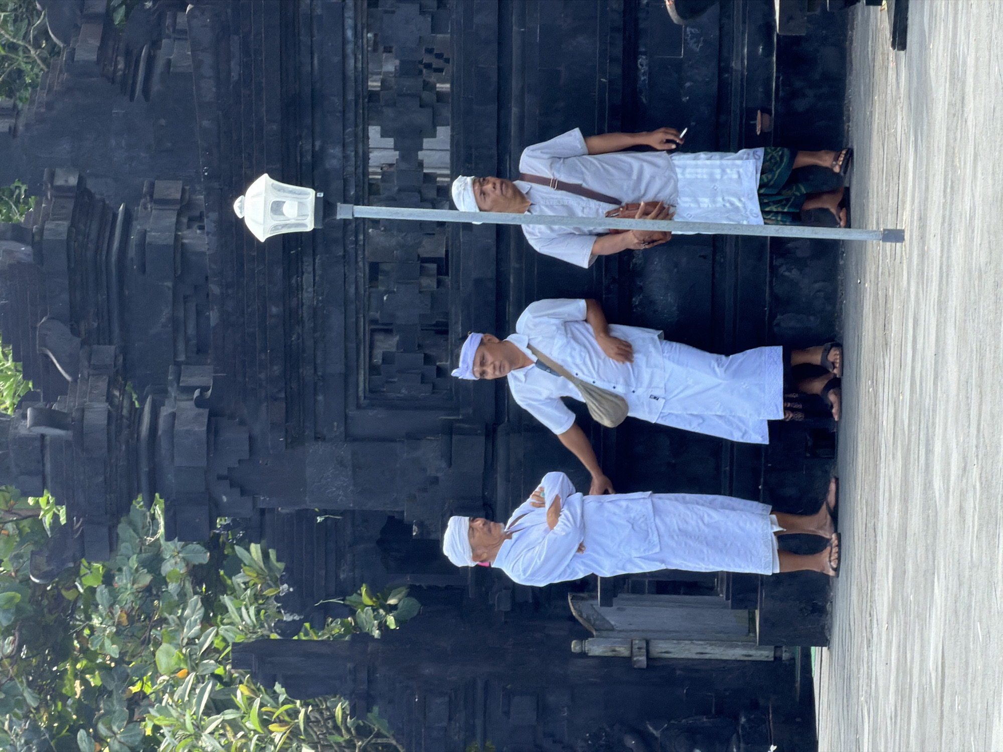 Three men are standing near a black stone structure with intricate carvings in Mengwi, Indonesia. The man on the left is wearing a white robe and headdress and has his arms crossed. The man in the middle is wearing a white shirt and sarong and has a tan bag slung across his chest. The man on the right is wearing a light gray shirt and a patterned sarong and has a brown bag across his chest, and he is holding a cigarette. A street lamp stands between the two men on the right. The background shows lush green foliage and a dark, ornate stone building.
