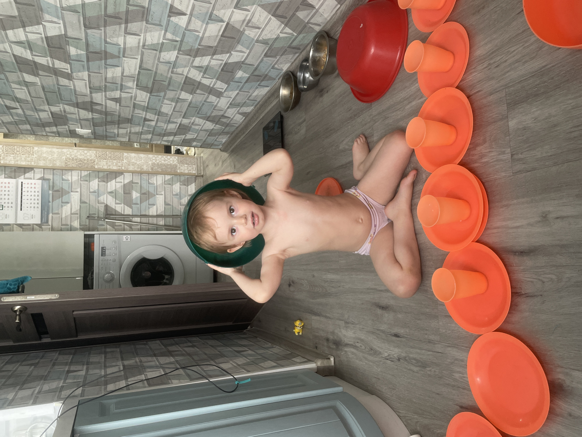 A young child is lying on a wooden floor, wearing a diaper. They are holding a green bowl over their head. The child is in the middle of a circular arrangement of orange plates with cups on them. To the child's left are several metal bowls and a large red bowl. In the background, a washing machine is visible next to a calendar. The walls are covered in textured wallpaper. The lighting suggests it is daytime. The child appears to be playing.