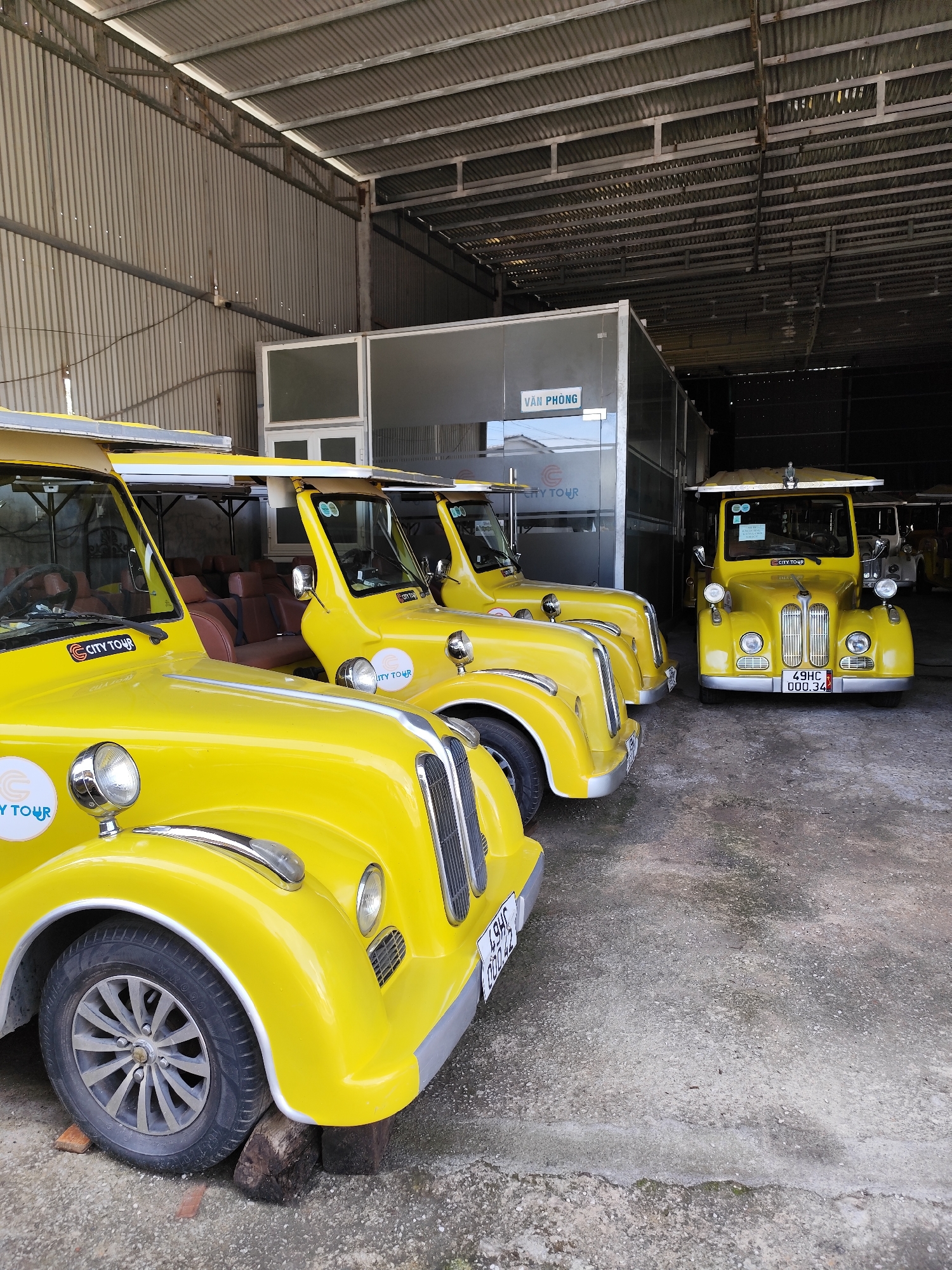 The image shows a collection of yellow "City Tour" vehicles parked inside a large, warehouse-like structure, likely a garage or storage area. Four vehicles are fully visible, with the front one most prominently displayed. The vehicles are stylized with a retro design. The "City Tour" logo is visible on the front of each vehicle. The vehicles' license plates are partially visible, with one displaying "49HC 000.34" and another displaying "49HC 000.42." The front vehicle's tire is positioned on top of wooden blocks. A structure labeled "VĂN PHÒNG" (Office) in Vietnamese is visible in the background. The corrugated metal construction of the warehouse is evident, with natural light entering the space. The vehicles likely offer tourist transportation. The "49HC" portion of the license plate suggests that the location is in the province of Lâm Đồng, where Đà Lạt is located. Thus, the image depicts vehicles in Xuân Trường – Đà Lạt, Vietnam.