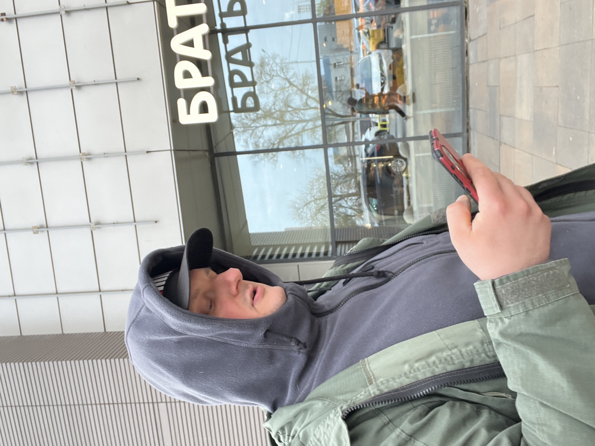 The image, oriented correctly, captures a man from the chest up, looking down at a red-cased smartphone he holds in his right hand. He is wearing a dark grey hooded sweatshirt with the hood up, underneath an olive green jacket, and a dark baseball cap is visible under his hood. His mouth is slightly open, suggesting he might be speaking or deeply engrossed.
The background features the modern facade of a building. Large white Cyrillic letters spelling "БРАТ" (BRAT, meaning "brother") are prominently displayed on the upper part of the building. Below this sign, a large window reflects the surrounding environment, including bare-branched trees (indicating late winter or early spring), cars, and a street scene. The reflection also shows an older spelling of the word, "БРАТЪ". The lower section of the building directly behind the man has a textured surface with thin vertical lines.
The scene takes place outdoors during the daytime in an urban setting, likely in Mytishchi, Russia, given the provided context. The weather appears to be cool or overcast. The man's focus on his phone is the central activity.