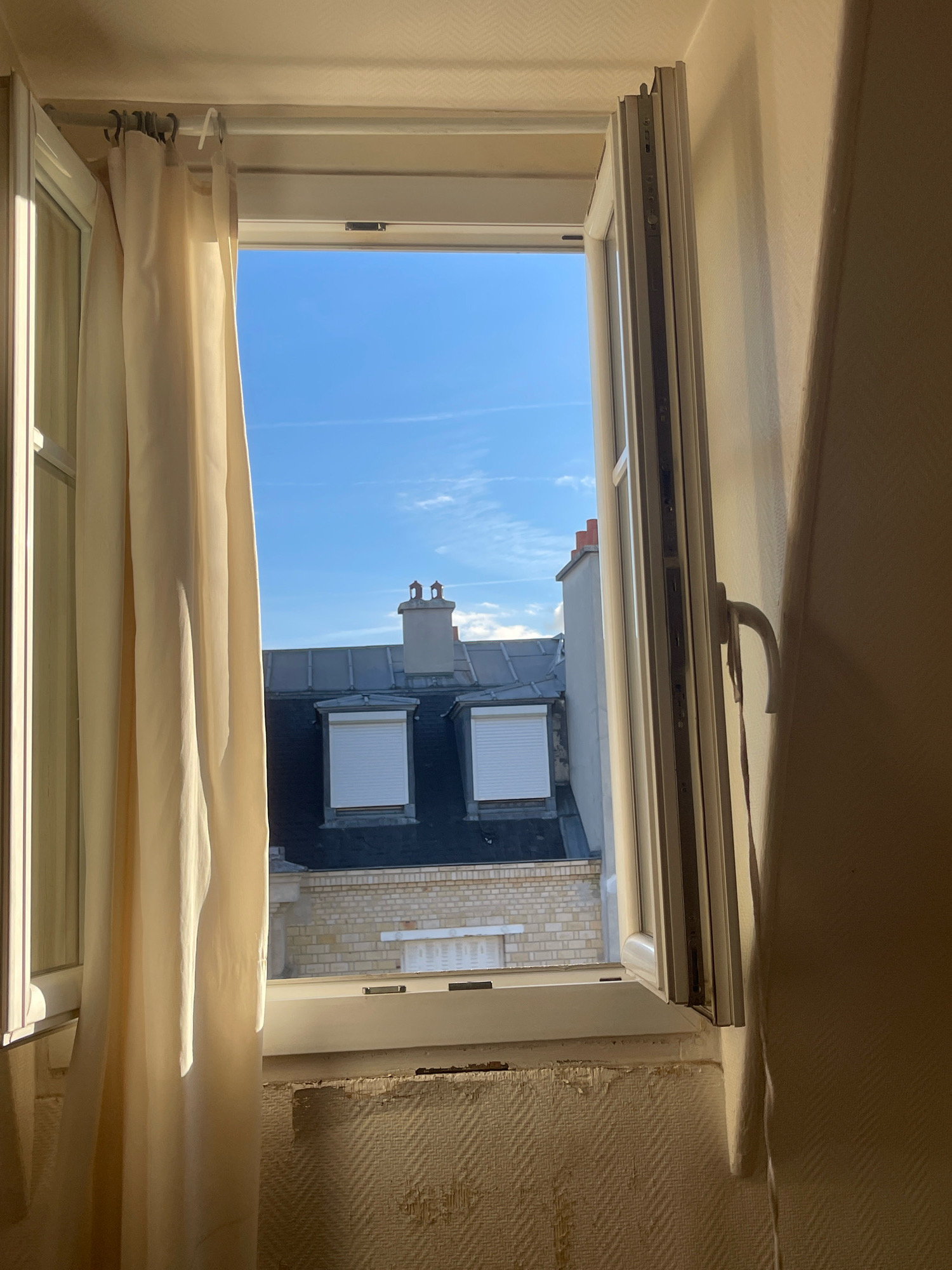 The image depicts an interior view looking out of an open window onto a Parisian cityscape. The window frame is white, and a light-colored curtain hangs on the left side, partially obscuring the view. Through the window, one can see the rooftops of buildings, characterized by dark gray slate roofs and chimneys. Two dormer windows with white shutters are visible. The sky is a clear blue with a few faint contrails. The interior wall below the window is covered with textured wallpaper, showing signs of wear and peeling in places. The lighting suggests it is daytime, with sunlight casting shadows. There are no people or animals present in the scene.