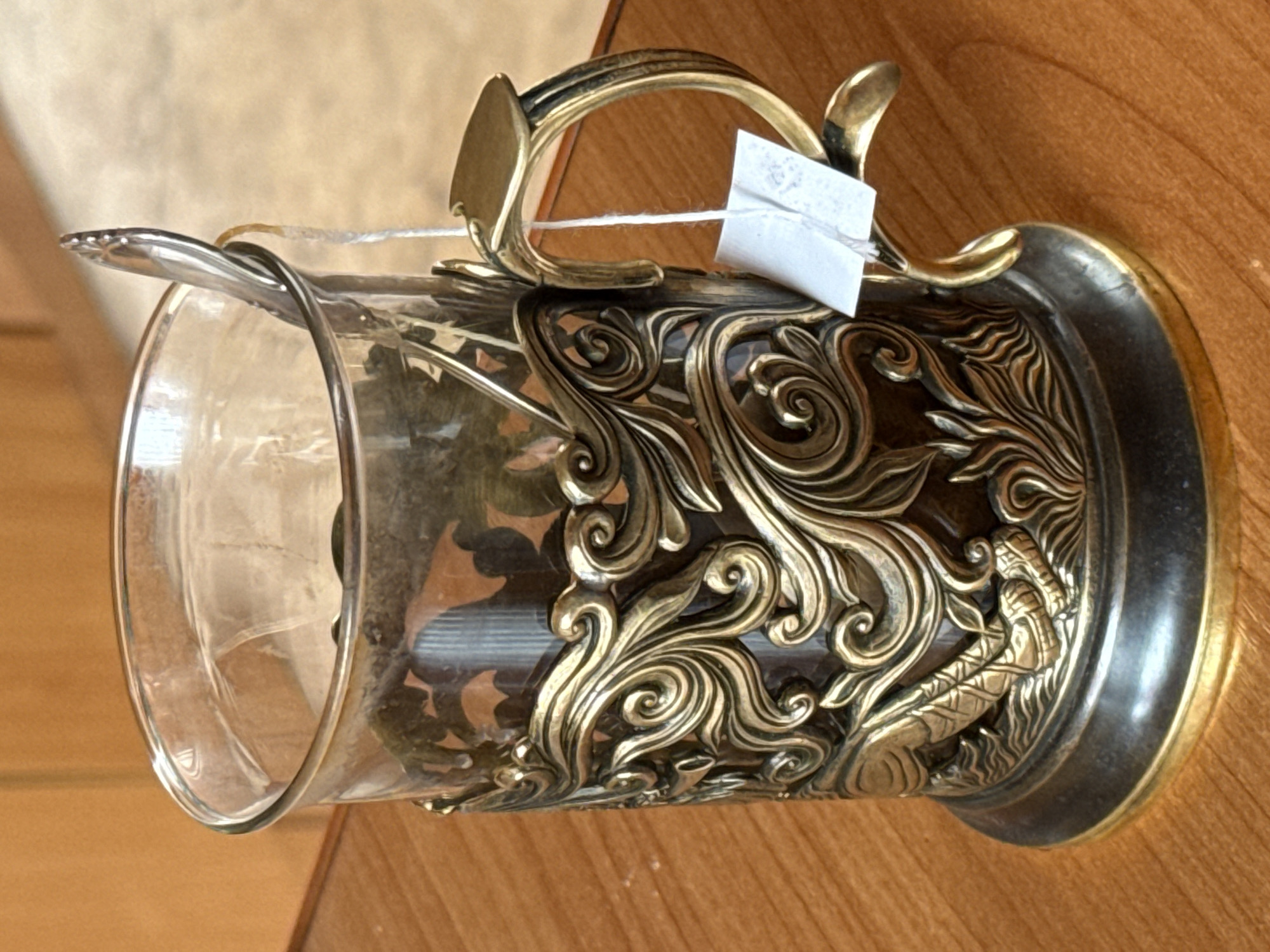 A close-up, eye-level shot shows a clear glass teacup held in an ornate brass holder. The holder is detailed with swirling patterns, and at the bottom, a relief of what appears to be a stylized fish or serpent. A small, white tag is tied with white string to the handle of the brass holder. A silver spoon is visible inside the glass, resting against the rim. The teacup and holder are placed on a wooden surface, possibly a table. The background is blurred but suggests a wooden cabinet or wall. There are no people present in the image. The scene is indoors, in an environment that is not specified as a city.