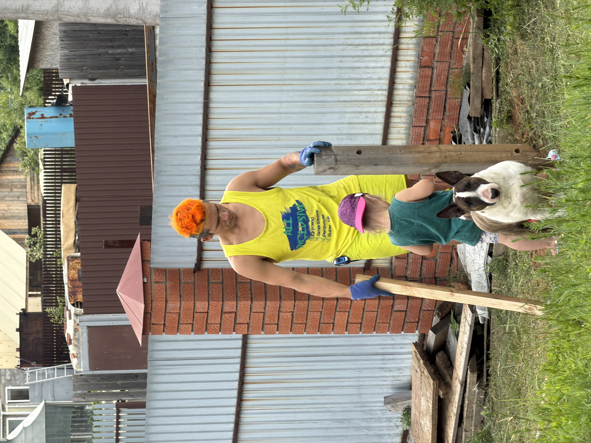 In Chaikovsky, Russia, a man with bright orange hair, wearing a yellow tank top and blue gloves, stands in a yard, holding a wooden plank. He is looking down at a young child wearing a green shirt and a pink hat, who is standing next to him, facing away from the camera. A bull terrier dog with a distinctive brown and white coat is positioned in front of the child, looking directly at the camera. The scene is set against a backdrop of a corrugated metal fence, a brick pillar, and piles of wood and construction materials, suggesting an outdoor work or renovation area. In the background, glimpses of buildings and greenery are visible, indicating a semi-urban or rural environment.
