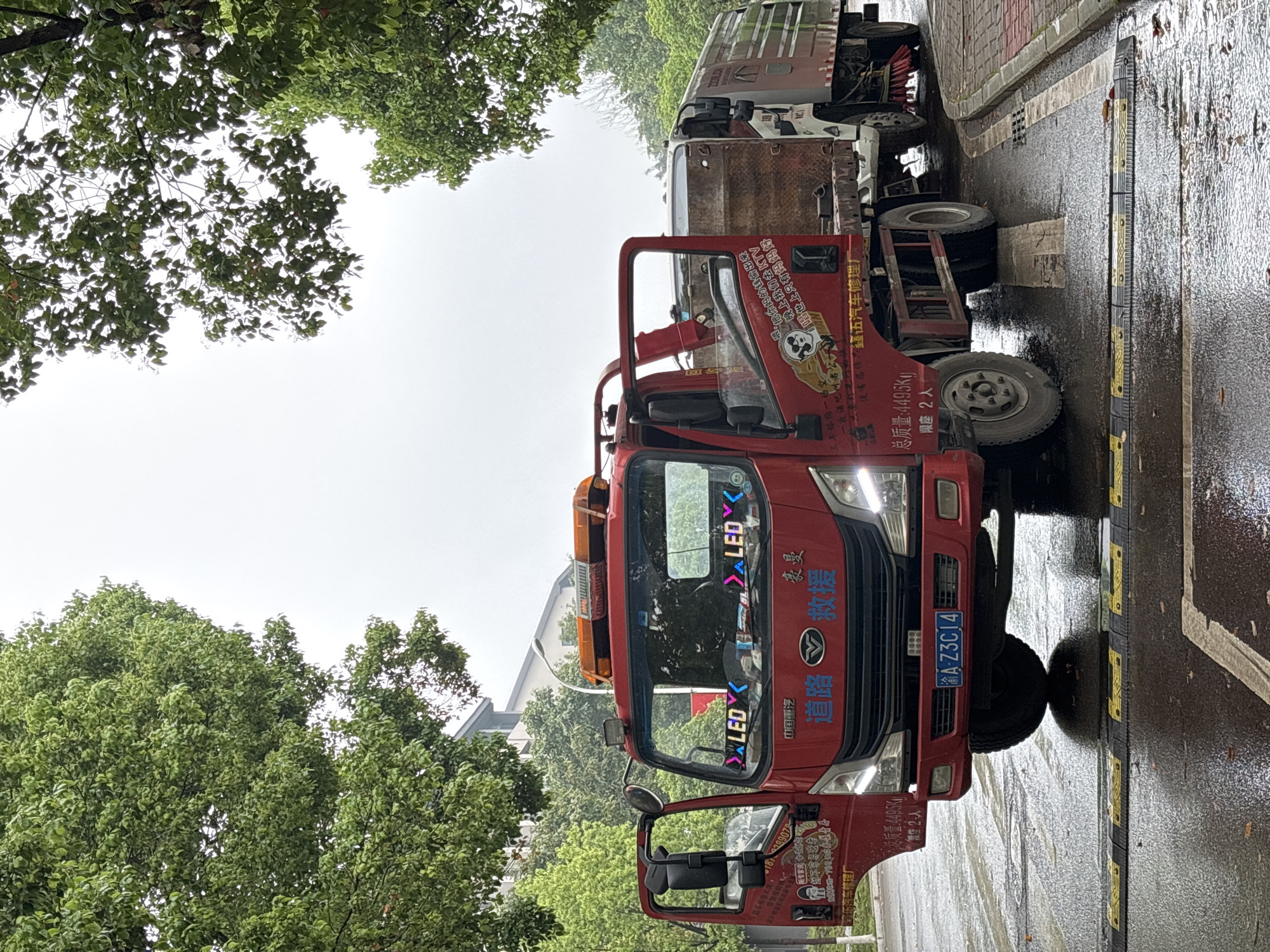 The image shows a red tow truck parked on a wet street, likely after a rain. The truck has Chinese characters and the license plate "渝A-Z3C14," suggesting it is located in Chongqing, specifically the Jiangjin district. Speed bumps are visible on the street. The truck is adorned with stickers and lettering in Chinese. The surroundings include trees and a building in the background. The OCR identified text includes "道路救援" indicating road assistance, "中国重汽" indicating China National Heavy Duty Truck Group, "豪曼" indicating Howo, a Chinese brand, and "江津伍汽车修理厂" possibly indicating a local car repair shop in Jiangjin.