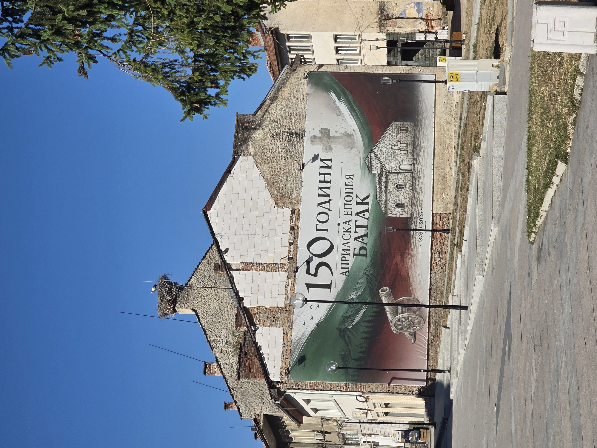 A bright, sunny day in Batak, Bulgaria, is captured in this image, highlighting a large banner draped across the facade of a weathered, multi-story building. The banner commemorates "150 ГОДИНИ АПРИЛСКА ЕПОПЕЯ БАТАК" (150 Years of the April Uprising Batak). It features a depiction of a historic church, a cannon, and a mountainous landscape with stylized trees and birds. A cross is also visible on the banner.
The building itself shows signs of age and repair, with exposed brick and stone elements. Above the banner, on the rooftop, a stork has built a nest on a chimney, and a stork is visible in the nest. The sky is a clear, vibrant blue. In the foreground, a paved plaza with tiered steps leads up to the building. Streetlights with spherical lamps are positioned along the plaza. A large pine tree partially obscures the right side of the frame. Electrical boxes and some vegetation are also present in the scene. No people are clearly visible or identifiable.