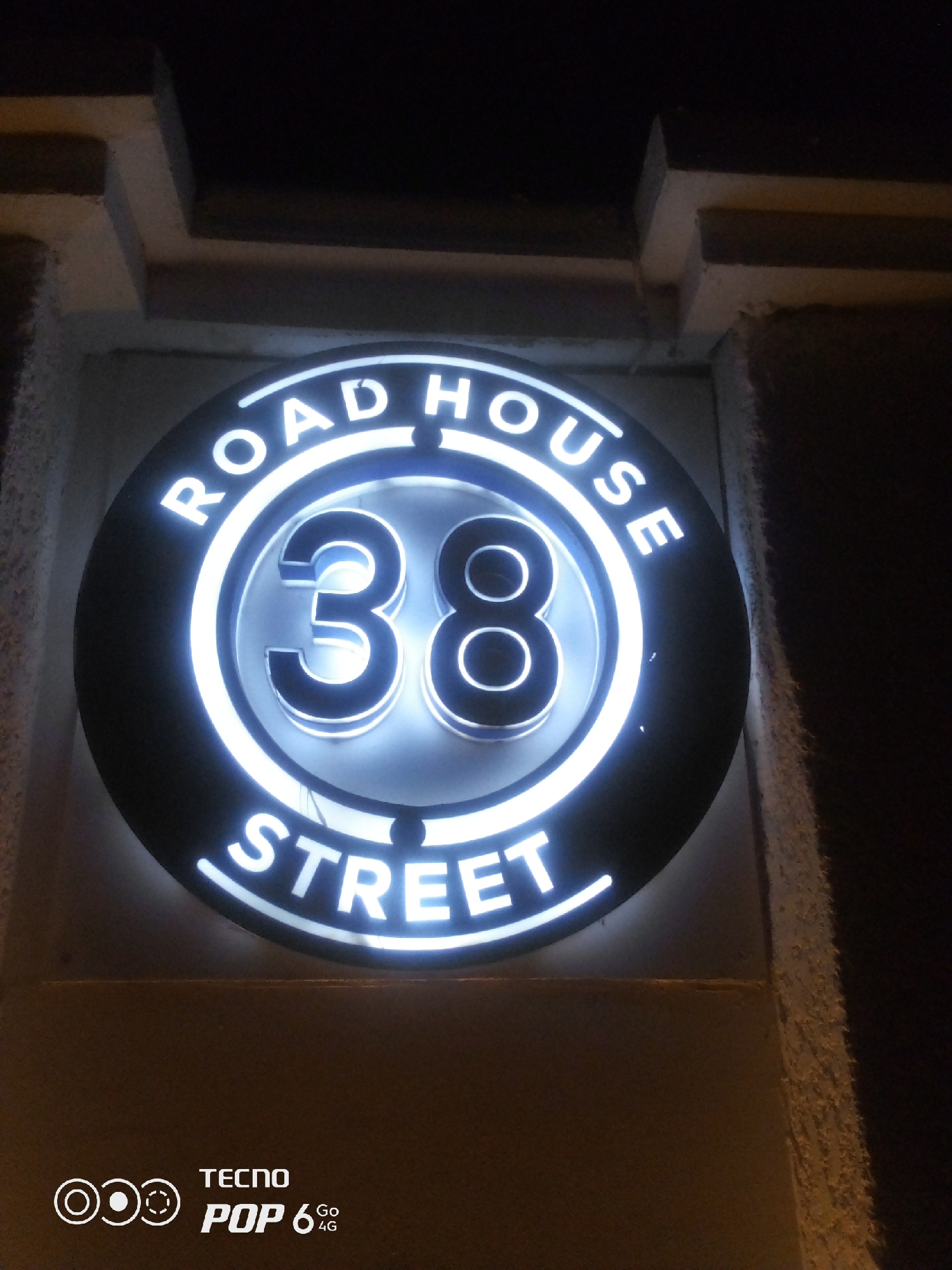 A circular illuminated sign reads "ROAD HOUSE 38 STREET". The sign is mounted on a textured, light-colored wall. Above the sign, there are architectural elements suggestive of a building's exterior, possibly a facade or overhang. The lighting from the sign creates a bright glow, illuminating the immediate surroundings. The bottom of the image displays a white logo and text that reads "TECNO POP 6 Go 4G". This is a street sign, likely indicating a business or residence located in Howayi, Nigeria.