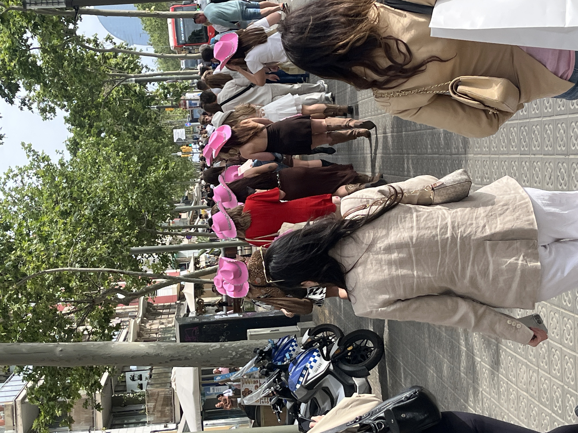 The image captures an outdoor urban scene in Barcelona, Spain, featuring a procession or gathering of people, predominantly women. Many participants are distinctively wearing bright pink cowboy hats, suggesting a themed event such as a bachelorette party or a public celebration.
In the foreground, several women are viewed from behind. One wears a beige blazer and carries a beige chain-strap handbag. Another wears a light beige jacket and holds a smartphone. The line of people extends into the mid-ground, showcasing various outfits including brown, red, and white dresses, blazers, and casual wear, all accessorized with the prominent pink hats.
The setting includes a tree-lined street with lush green foliage, urban buildings in the background, and a characteristic grey patterned pavement. Traffic lights and lampposts are visible. In the mid-ground, two scooters or motorcycles are parked, one blue and white with the visible text "GU-239". A red bus with partially legible text, including "PL CLIMENT 8-9A", can be seen in the distance. The scene is brightly illuminated, indicating daytime and clear, sunny weather. The atmosphere appears lively and communal.