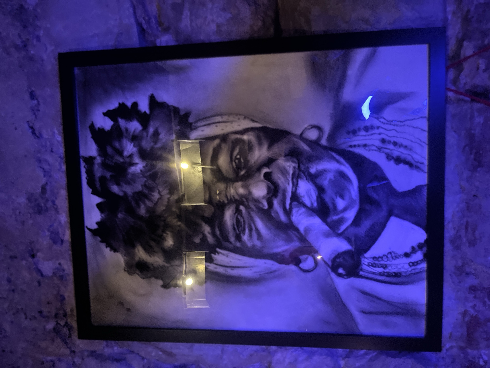A black-framed artwork hangs on a wall with textured, stone-like appearance. The artwork is a grayscale image of a person with dark hair, a bandana, and a cigar in their mouth. Reflective light sources are visible on the artwork's surface. The environment is dimly lit with a blue hue. The location is not apparent from the image.