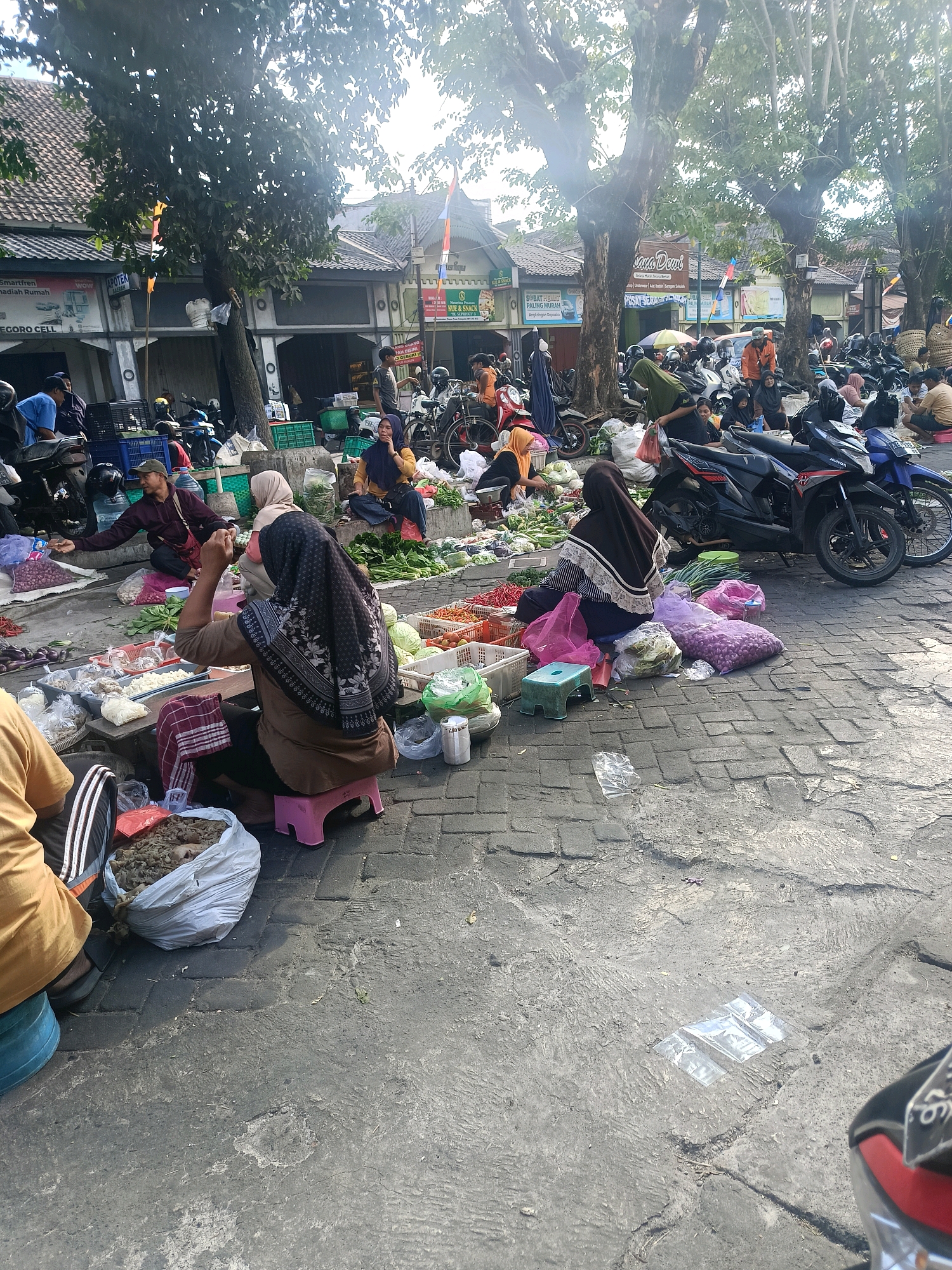 In Yogyakarta, Indonesia, a bustling outdoor market is captured under the shade of large trees. The scene is a vibrant display of commerce, with vendors and shoppers engaged in transactions. A variety of fresh produce, including vegetables and what appear to be small fruits or spices, are laid out on mats and in baskets. Several individuals, predominantly women wearing hijabs, are seated or kneeling, attending to their goods. A woman in a brown hijab and dress is seated on a pink stool, her hands clasped in front of her as she looks towards her offerings. Another woman, also in a hijab, is seated on the ground, sorting through produce. In the background, motorcycles are parked, suggesting the presence of customers or vendors arriving by vehicle. Buildings with traditional Indonesian architecture line the street behind the market stalls, some displaying signage for businesses like "APOTEK" (pharmacy). The ground is paved with cobblestones, characteristic of many older urban areas. The atmosphere suggests a typical day at a local market, with the natural light filtering through the leaves of the trees creating a dappled effect.