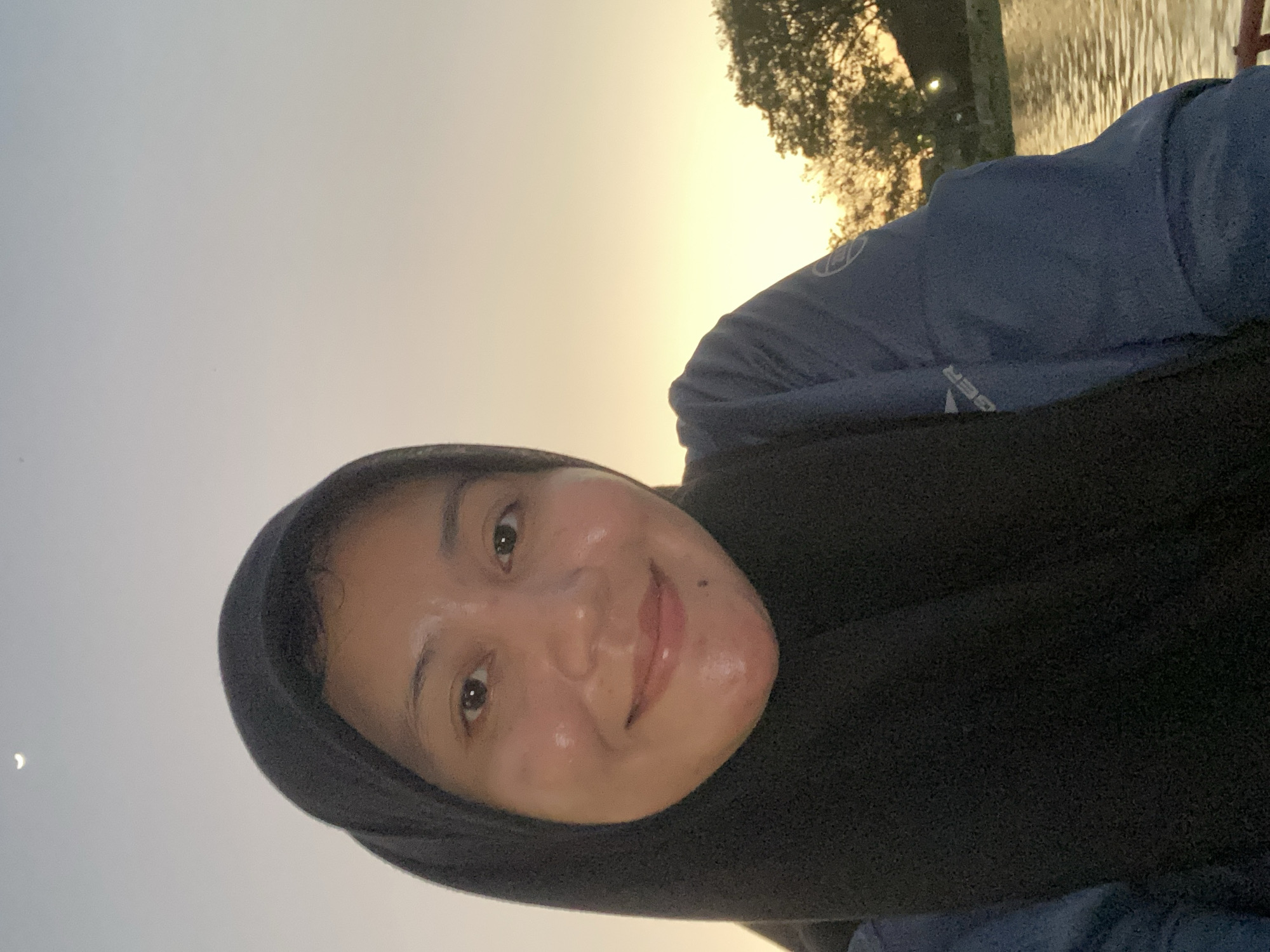 This is a close-up selfie of a woman wearing a black hijab. She is looking at the camera and smiling. The background shows a body of water, trees, and a warm sky, suggesting it is either sunrise or sunset. The location is identified as Banjarmasin, Indonesia. She is wearing a dark blue long-sleeved shirt. There is a crescent moon visible in the upper left corner of the sky.