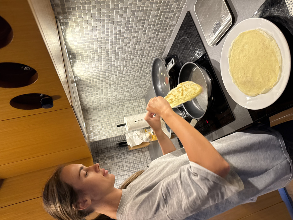 A female subject is captured in profile, mid-action, cooking in a contemporary kitchen located in Warsaw, Poland. She is seen from the waist up, dressed in a grey t-shirt, holding a cooked crepe or pancake with both hands, positioning it over a black frying pan situated on an induction stovetop. A second frying pan is present on the stovetop, and a finished crepe rests on a white plate to the right. The kitchen environment features dark grey countertops, a light grey mosaic tiled backsplash, and distinctive light wooden upper cabinets with circular cutouts. A stainless steel range hood, equipped with lights, is installed above the cooking area. Essential objects on the counter include a paper towel roll on a stand and a silver digital kitchen scale. The scene depicts domestic culinary activity.