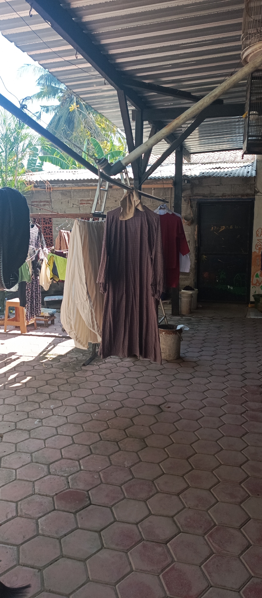 This is a sunlit, outdoor scene in Arjasa, Indonesia, featuring laundry drying under a corrugated metal roof supported by a metal frame. In the foreground, a patterned hexagonal paver stone patio is visible, with shadows cast by the laundry and roof structure. The patio is partially covered by a dark object in the lower left corner. Hanging on a clothesline are several garments, including a dark patterned dress on the left, a cream-colored sheet or fabric, and a dark brown patterned dress with a tan scarf draped over it in the center. To the right of the brown dress, a red shirt is hanging, and behind it, a white garment. In the mid-ground, a brick wall with a decorative screen is visible, along with a metal ladder leaning against it. Further back, a dark door with some writing and graffiti is present, and to the right of the door, a birdcage hangs from a pole. Tropical foliage, including palm and banana leaves, can be seen in the background, suggesting a warm climate. Several buckets are placed on the ground, some appearing weathered. The scene depicts a typical domestic setup for drying clothes in a residential or semi-rural area.