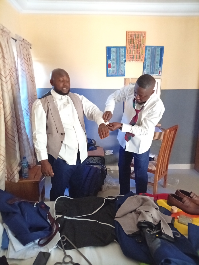 Two men are in a room, preparing. One man, wearing a vest and dress pants, stands while the other, wearing a white dress shirt, jeans, and a tie, adjusts his cuff. The room is decorated with artwork and has a small table with a water bottle. Clothes are scattered on a bed in the foreground, with shoes placed on top.