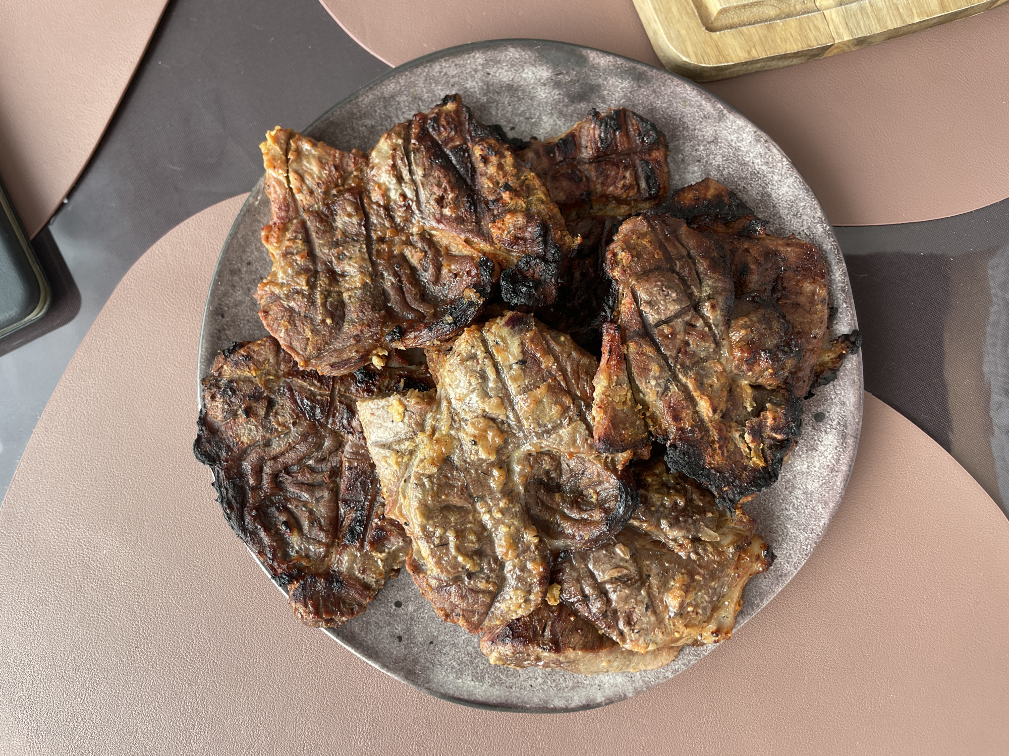 A top-down shot shows a gray, textured plate laden with several pieces of grilled meat, possibly pork or beef, characterized by prominent grill marks and varying degrees of charring. The meat appears to have been seasoned, evidenced by visible herbs and spices. The plate rests on a multi-toned place setting of brown and tan on a table. A dark gray cell phone is partially visible at the left, and a wooden cutting board is visible on the top right. The image was taken in Подшивалово, Russia.