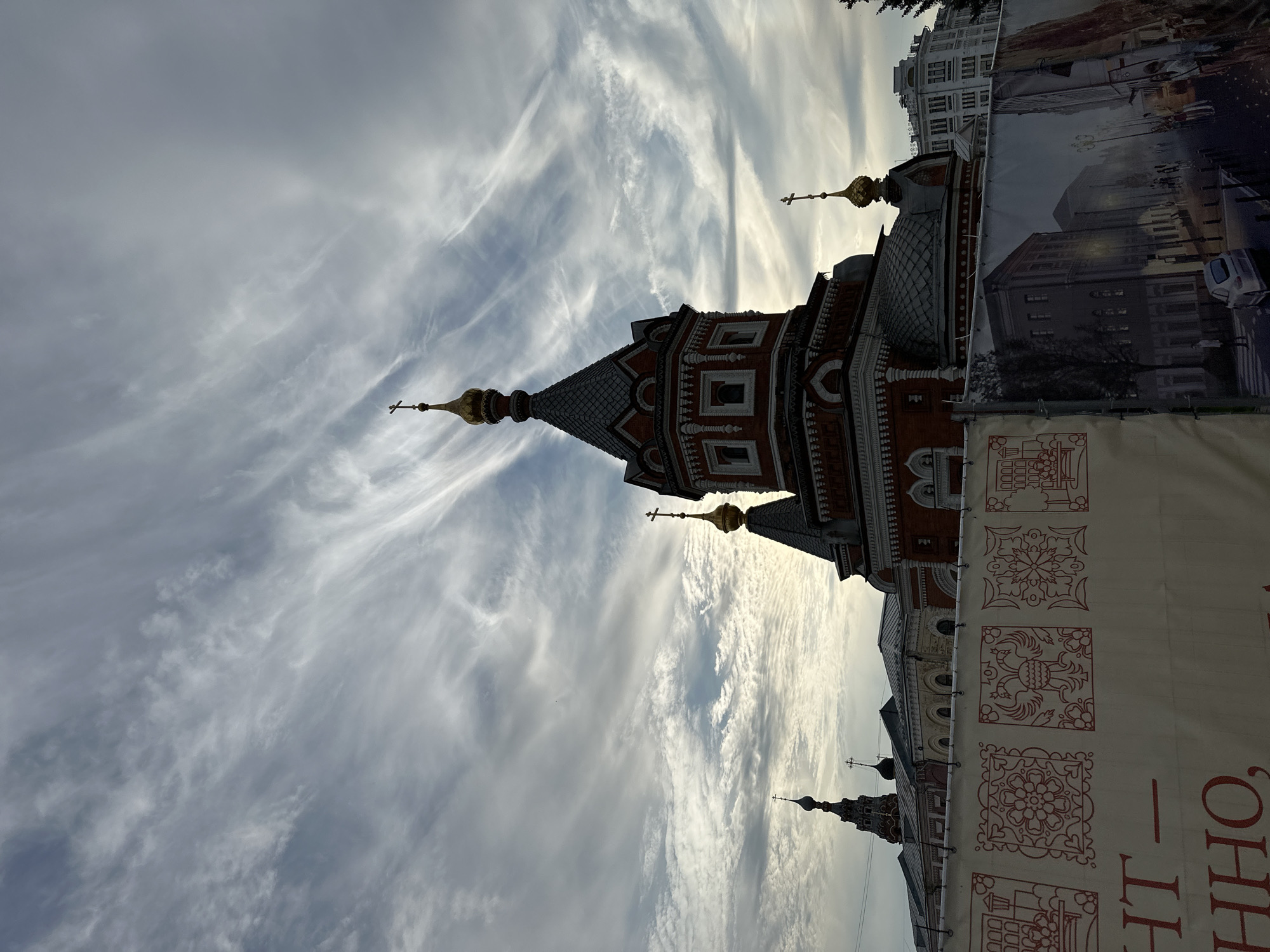 The image shows an Orthodox church with multiple domes, silhouetted against a cloudy sky. A banner with decorative patterns and text in Cyrillic characters is in the foreground, obscuring the lower portion of the church and surrounding buildings. The banner also features a printed image of a street scene with buildings and people. The overall impression is that of an urban environment, possibly undergoing some form of construction or renovation given the presence of the banner. The location is identified as Putyatino (Semivragovskaya S/A), Russia.