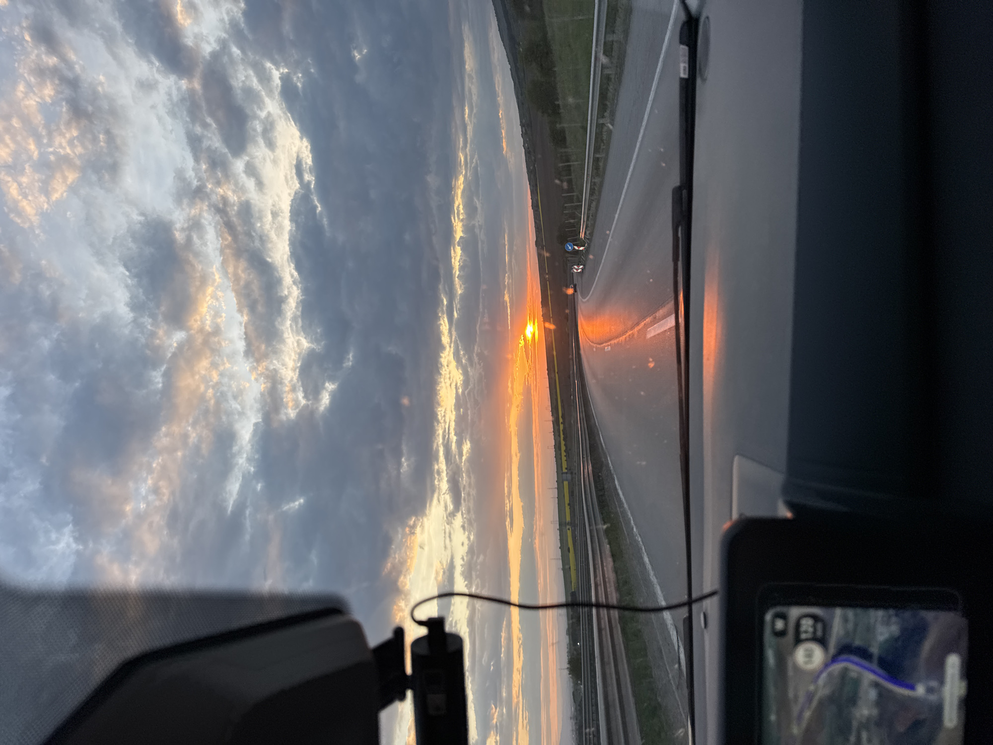 The image, taken from inside a vehicle, shows a sunset over a highway. The sun is low on the horizon, casting orange light across the road and sky. Dark, gray clouds fill the sky above the sunset. The road is marked with white lines and a yellow median. Fields are visible on either side of the highway. A navigation device is visible in the lower right corner of the image, displaying a map with a blue route. The map shows a section of a road with a curve or bend. Although the navigation device may indicate a route that potentially involves the city of Eleshnitsa, Bulgaria, no details within the image explicitly confirm the location.