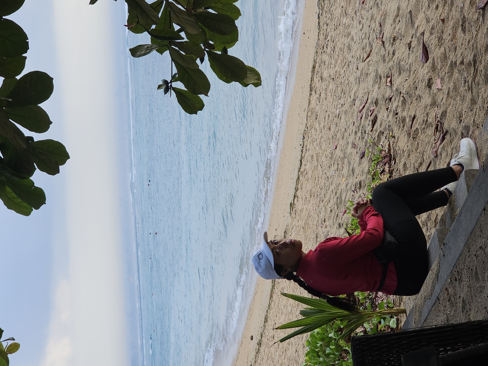 A woman wearing a red long-sleeved shirt, black leggings, and a white baseball cap sits on a bench facing the ocean on a sandy beach in Denpasar, Indonesia. She is looking to her right, away from the camera, with her hands clasped in her lap. Green leaves from a tree hang into the frame from the top, obscuring part of the sky. The ocean is calm with small waves breaking on the shore, and there are buoys visible in the distance. The sand is littered with dry leaves.