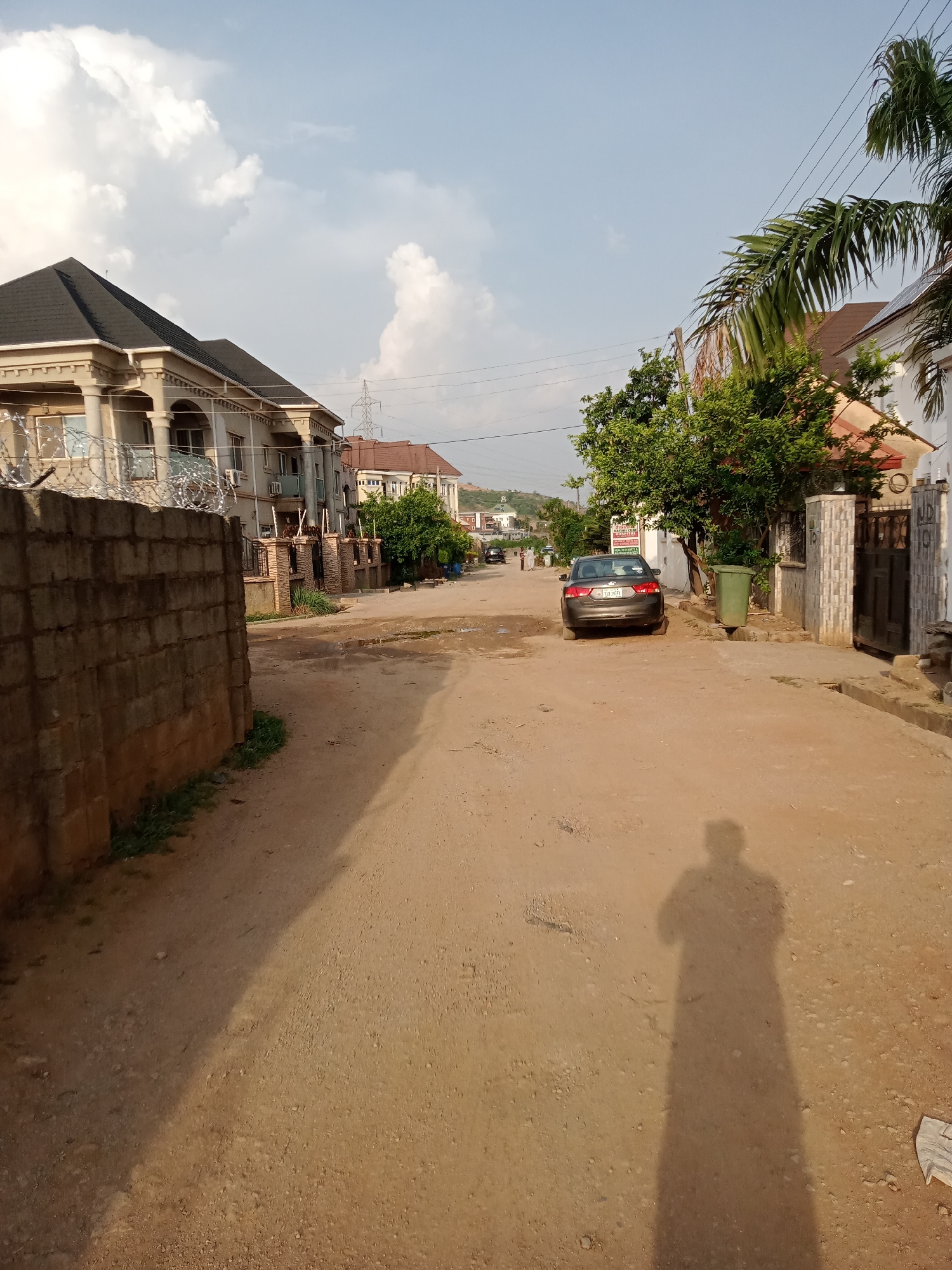 The image shows a street in Gwarinpa, Nigeria. The road is unpaved with a dirt surface. A dark gray sedan is parked on the right side of the street. Buildings line both sides of the street, including a two-story building with a gray exterior on the left side which is surrounded by razor wire on top of a brick wall. There are power lines visible in the background. A green trash bin sits to the right side of the road. The shadow of a person taking the picture can be seen in the foreground. The sky is partially cloudy.