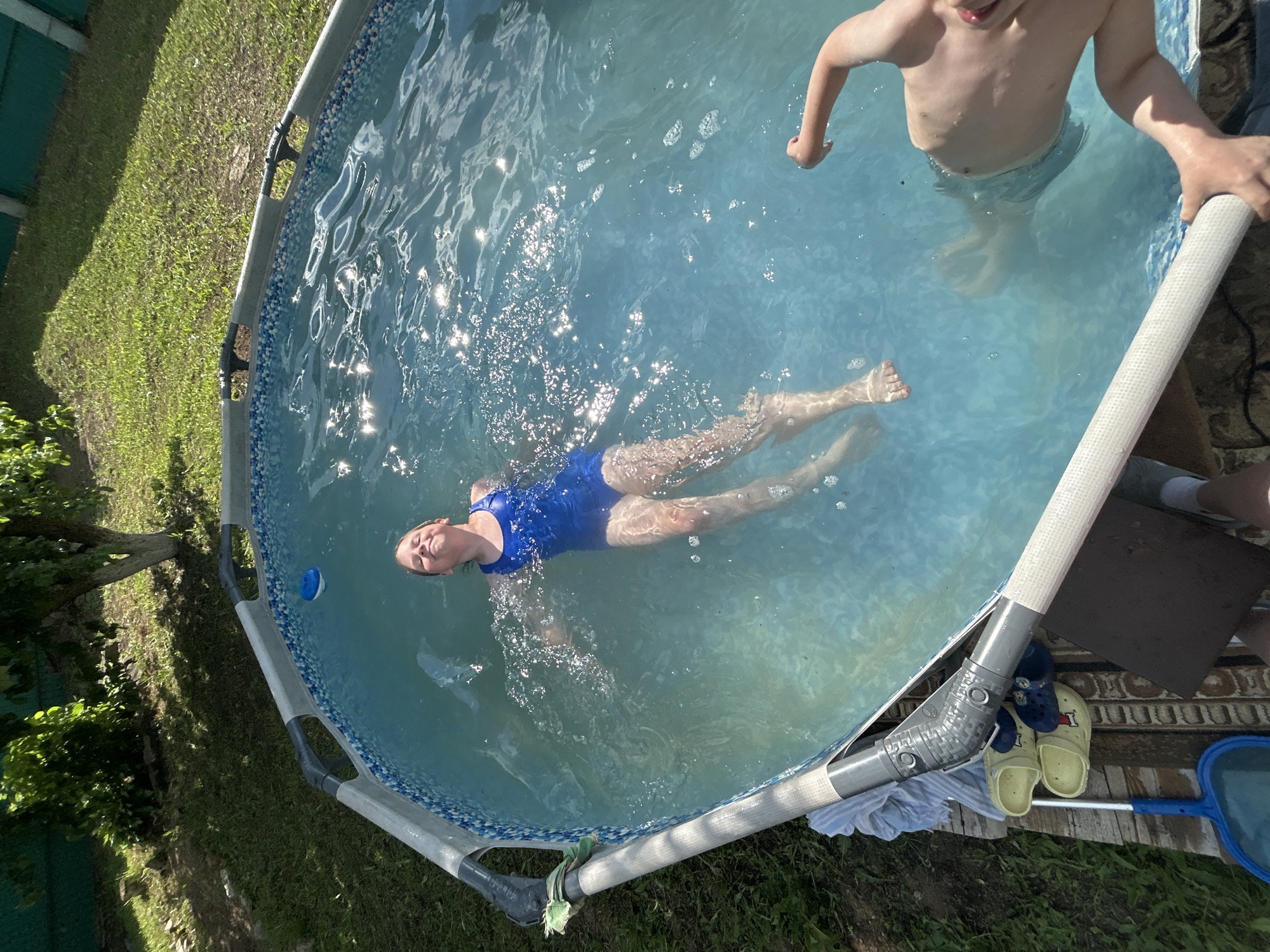 The image shows an above-ground swimming pool in a backyard. A young girl in a blue swimsuit is floating on her back in the water, smiling. Another child is partially visible in the lower right corner of the frame, with their arm extended. The pool is surrounded by grass and trees. There are also a pair of yellow and blue clogs and a blue pool skimmer visible on the ground next to the pool. The location is the city of Fokino, Russia, judging by the context provided by the user.