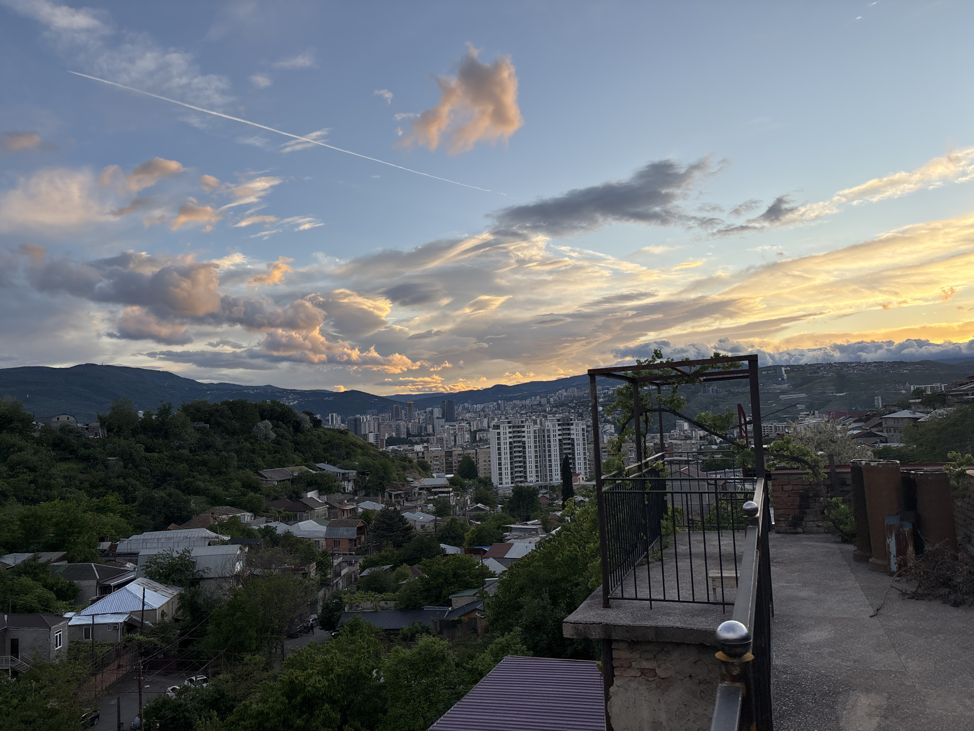 The image shows a cityscape view of Tbilisi, Georgia, under a cloudy sky with streaks of sunlight. A long contrail cuts across the upper left sky. A series of buildings are situated along a hill, with a concentration of taller structures forming a skyline in the central background. Mountains are faintly visible behind the buildings. In the foreground, a terrace with a metal railing and trellis framework overlooks the scene. A mix of residential houses and dense green trees fill the lower left area. The terrace surface is concrete and slopes down toward the edge. There are three metal, cylindrical containers on the right edge of the terrace. The time of day appears to be either early morning or late afternoon, given the soft, diffused light.