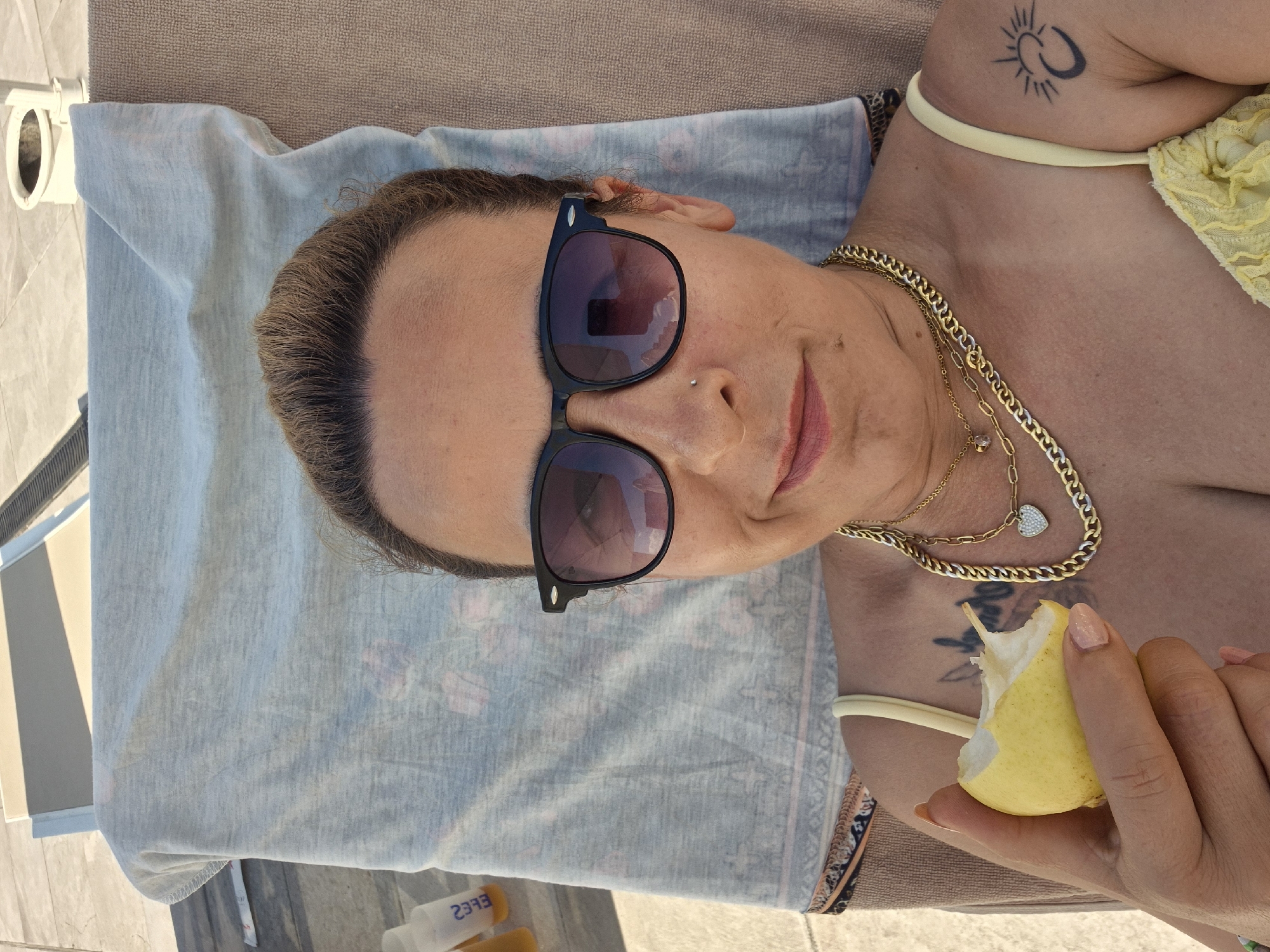 A woman is lying on a lounge chair on a sun-drenched patio, holding a bitten pear in her left hand. She is wearing sunglasses, a yellow bikini top, and multiple gold necklaces. A tattoo of a crescent moon and sun is visible on her right shoulder. The scene appears to be outdoors, possibly near a pool or beach, with the sun casting shadows.
