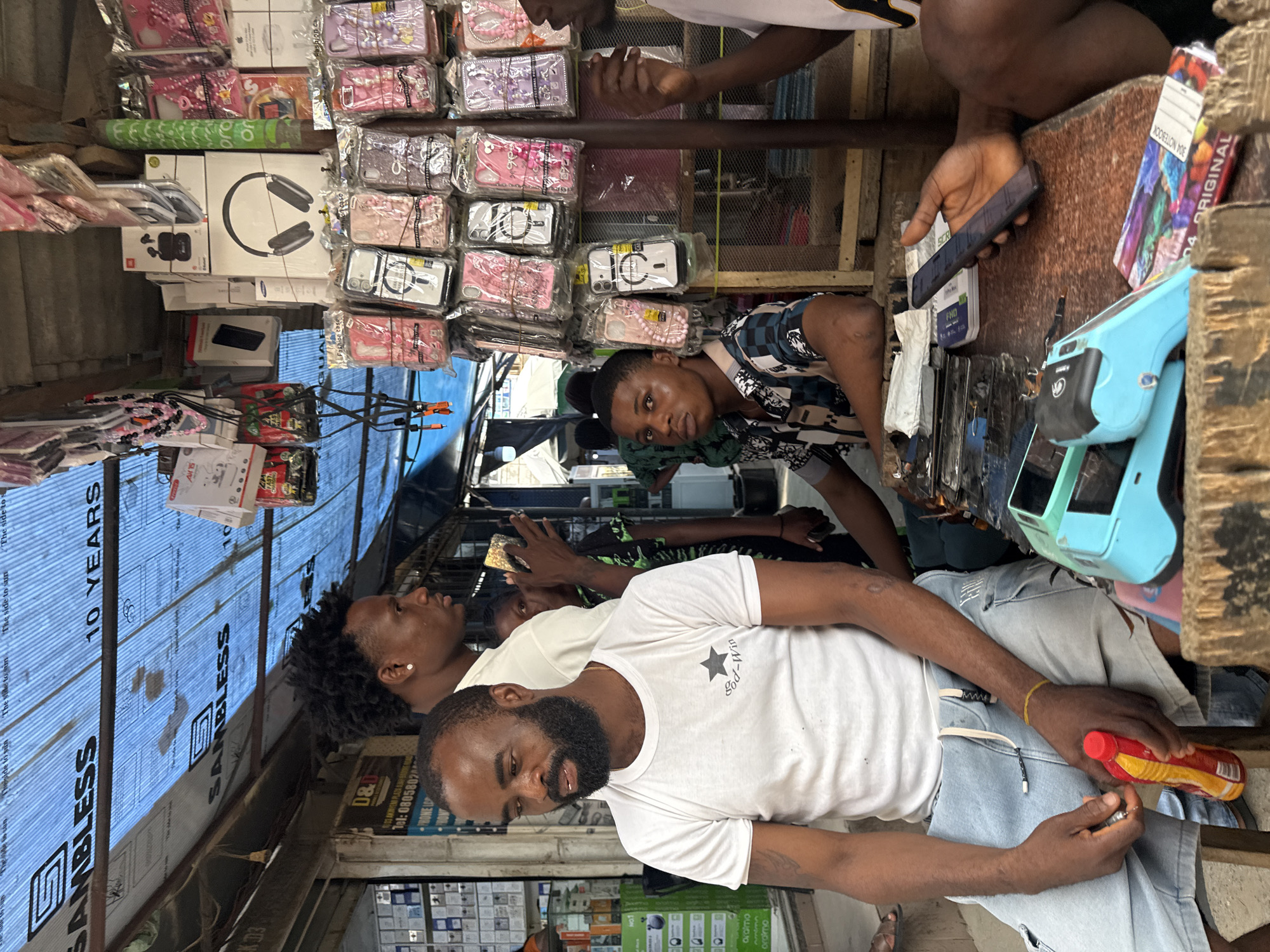 The image depicts a street market scene in Lekki, Nigeria, likely at a stall selling mobile phone accessories. In the foreground, a man with a full beard and dark skin sits on a wooden stool, wearing a white t-shirt with a star graphic and light blue denim shorts. He holds a red and yellow bottle and appears to be looking down. To his right, another young man with dark skin and a patterned short-sleeved shirt is seated, looking towards the left. Behind them, several other individuals are visible. The stall itself is covered by a blue tarpaulin with "SAMBLESS" branding visible. The stall is laden with various items, including stacks of mobile phone cases displayed on racks, boxed electronics like headphones and earbuds, and what appears to be a point-of-sale device and mobile phones on the counter. The overall environment suggests a bustling outdoor market with goods displayed for sale under a makeshift canopy.