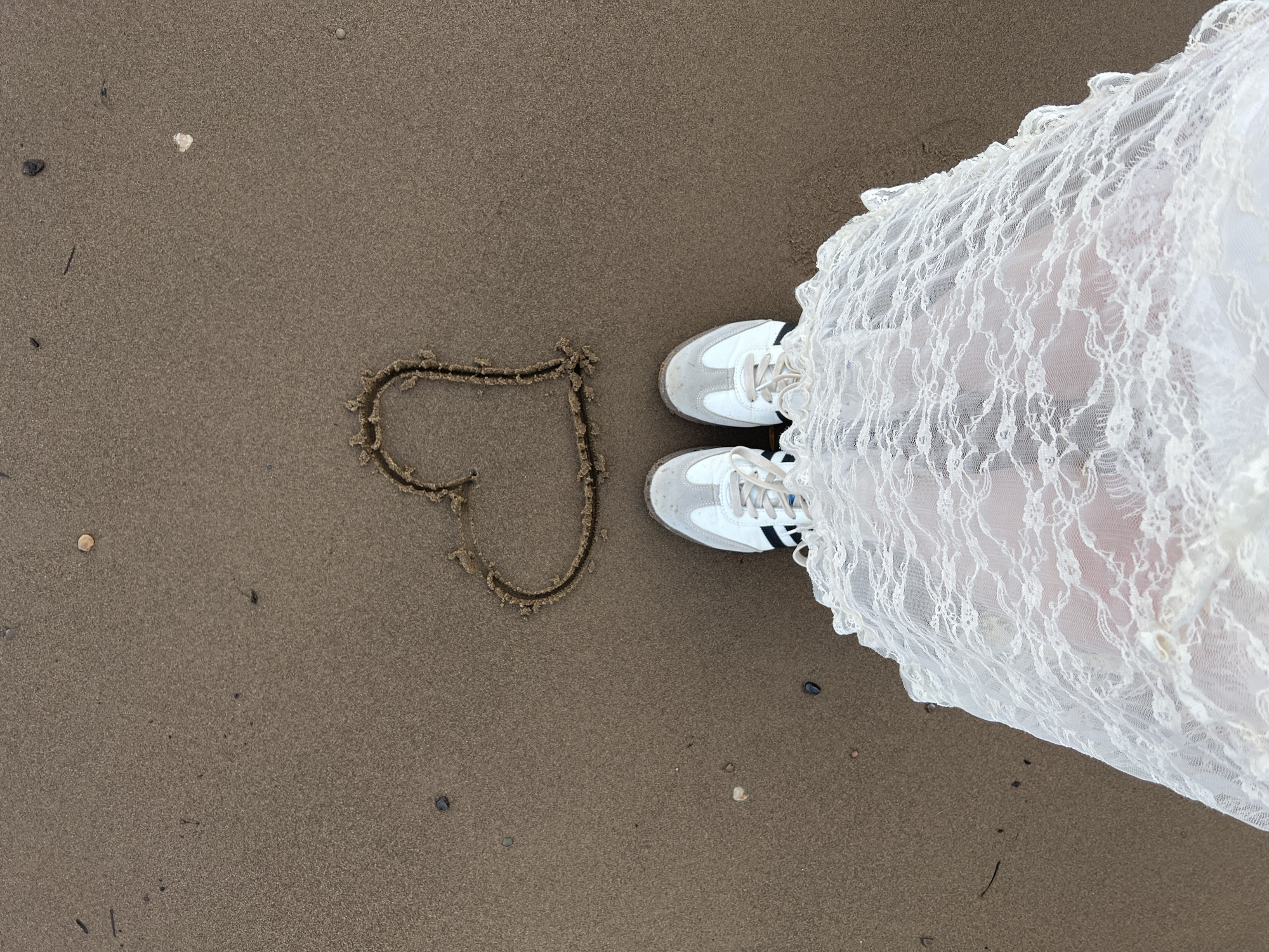 A pair of white and grey sneakers are standing on a sandy beach, in front of a white lace skirt that drapes over the person's legs and extends down. A heart shape has been drawn in the wet sand between the person's feet and the camera. Small stones and debris are scattered across the sand.