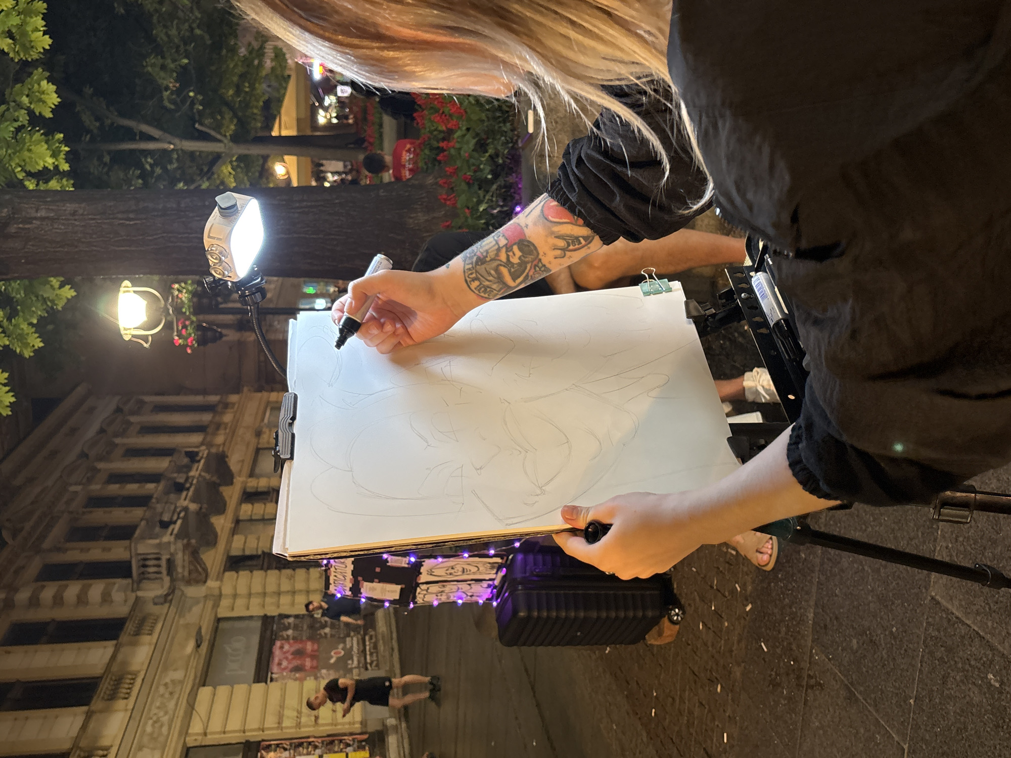A street artist with blonde hair is drawing a caricature on a large white paper using a black marker. The artist has a tattoo on their right forearm and is wearing a black shirt. A bright white light illuminates the drawing surface. The easel is set up on a cobblestone street. In the background, there is a building and people walking by. Purple lights are strung around the base of the drawing. The location is likely in Belgrade, Serbia, as there is a poster advertising "Karikature" at the location.