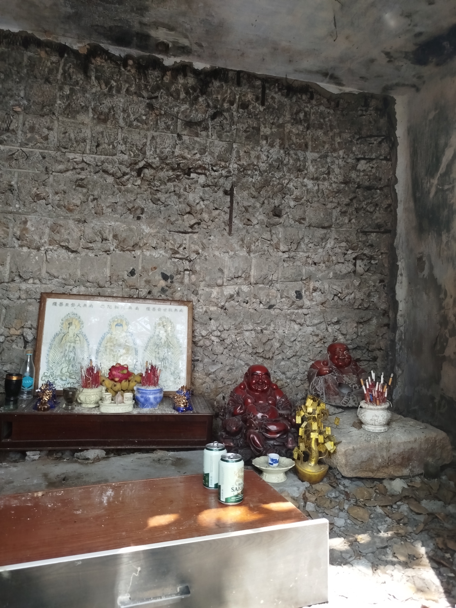 The photograph depicts an indoor shrine in what appears to be a dilapidated or unfinished room. The room is constructed with rough, grey stone walls and a bare ceiling.
At the back of the room, a wooden table serves as the main altar. A framed picture featuring three figures and some Vietnamese characters, along with offerings such as fruit and incense sticks in colorful ceramic bowls, sits prominently on the table. To the right, two laughing Buddha figurines are placed on a large stone.
In the foreground, a wooden table with a metal base holds two cans of "Saigon" beer. The floor around the table and altar is covered in debris, giving the impression of an abandoned or forgotten space.
Given the presence of "Saigon" beer, the location is likely in or around Ho Chi Minh City, Vietnam.