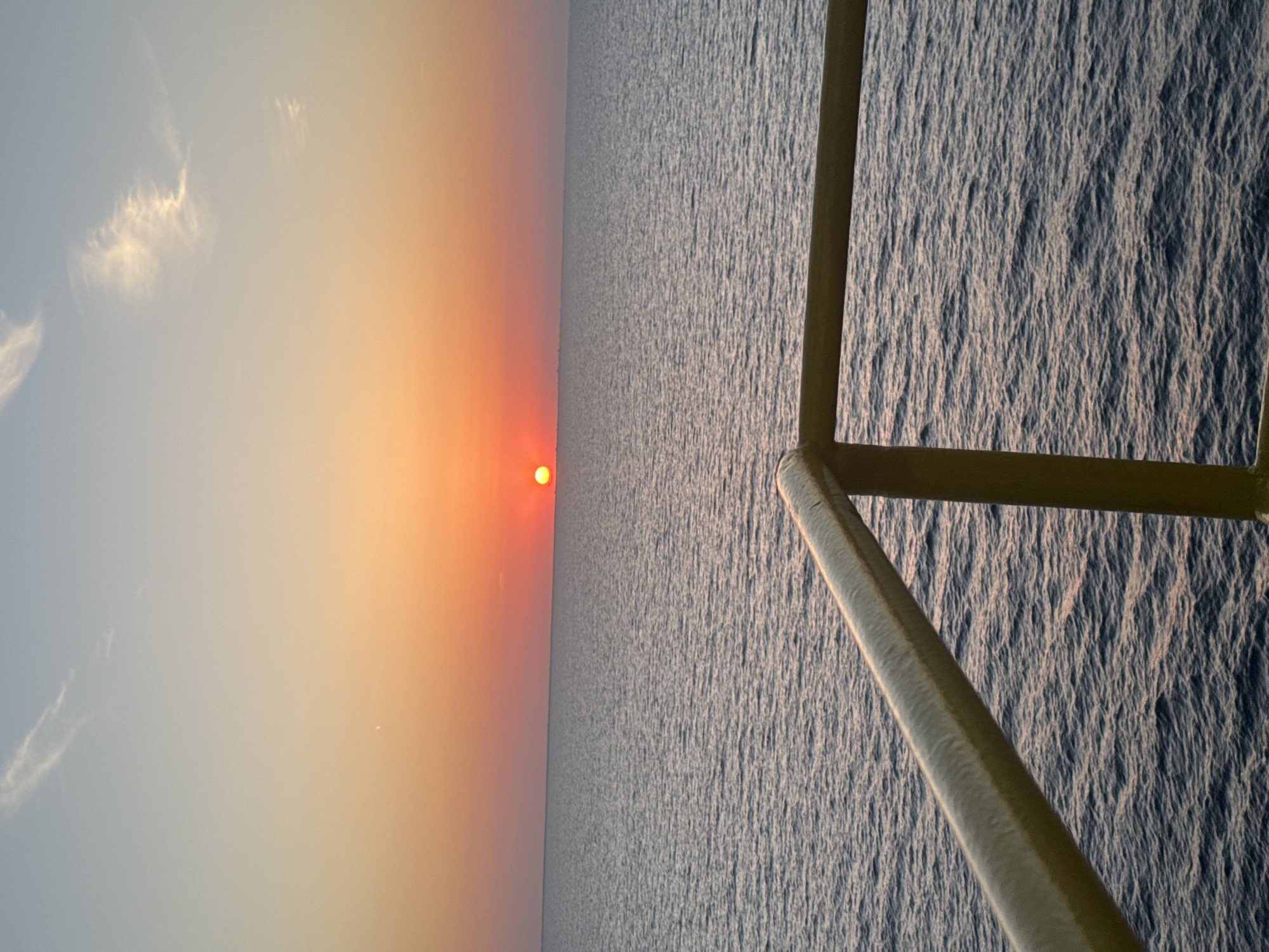The image captures a serene scene of the sun setting over a calm ocean. The sun, a vibrant orange orb, is low on the horizon, casting a warm glow across the sky and reflecting on the water. The sky transitions from a soft blue at the top to a gradient of orange and yellow near the horizon, with a few wispy clouds adding texture.
Dominating the right side of the frame is a metal structure, possibly part of a boat or a pier, with thick, rounded beams. These beams are a light tan color and appear to be made of metal, with some visible texture suggesting wear or a painted surface. The structure is angled, creating a diagonal line that divides the image and draws the viewer's eye towards the seascape.
The ocean's surface is textured with small ripples, indicative of gentle waves, and reflects the light of the setting sun, giving it a shimmering, silvery-blue hue. The horizon line is sharp and distinct, separating the sky and sea. The overall atmosphere is peaceful and tranquil, evoking a sense of calm and the beauty of nature. The time of day is clearly sunset, and the weather appears to be clear and fair. There is no visible text in the image.