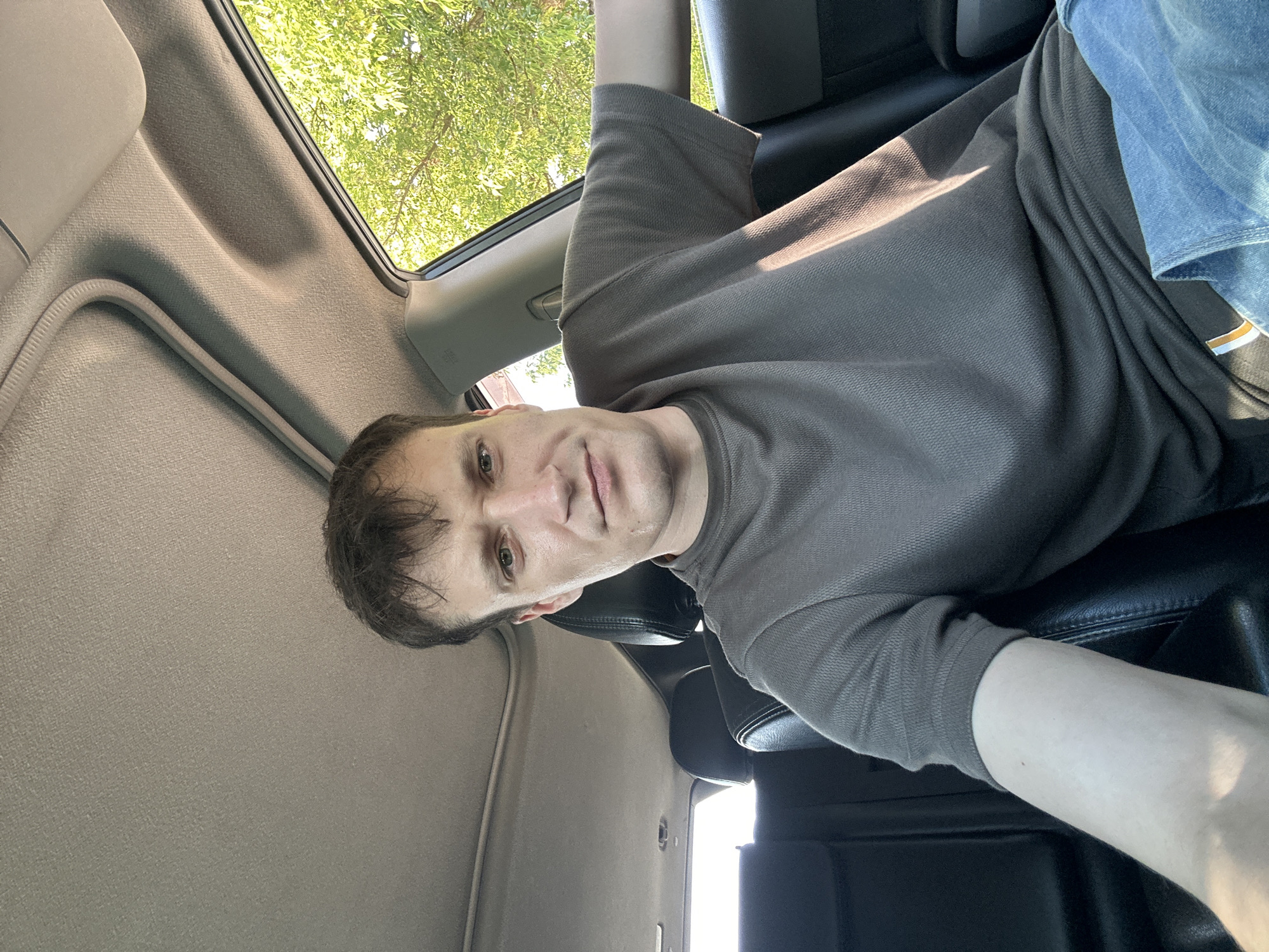 A male, appearing to be in his late 20s or early 30s, is seated in the back seat of a car. He has light skin, dark brown hair, and light green eyes. He is wearing a short-sleeved, grey polo shirt and blue jeans. The car's interior is visible, with black leather seats and a grey headliner. Outside the car, green trees and foliage are visible through the open window, suggesting an outdoor setting, possibly in Makiv, Ukraine. The image is taken from a selfie perspective, with the man looking directly at the camera.