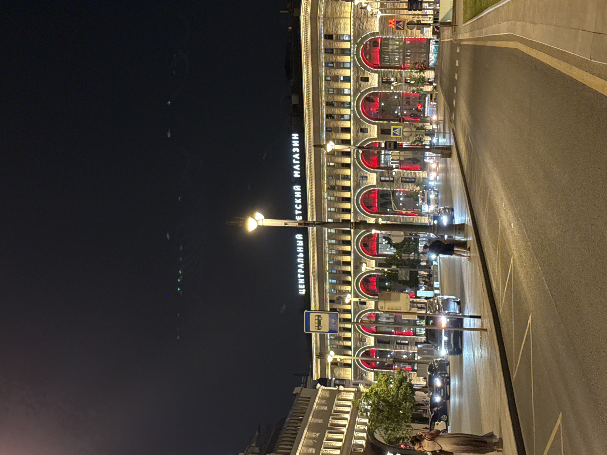 At night, a street scene in Moscow, Russia, shows illuminated architecture and a road surface. The building to the left, identifiable by the Cyrillic inscription "ЦЕНТРАЛЬНЫЙ ДЕТСКИЙ МАГАЗИН" ("Central Children's Store"), is brightly lit, featuring red arches and multiple windows. Streetlights illuminate the street and cast shadows of people and vehicles. A street with markings stretches along the right side of the scene. Cars are parked or moving along the street. A few people are visible in the foreground, including at least one pair who appear to be embracing. Signage, including bus stop indicators, is visible, along with some sparse greenery. The sky is dark, suggesting a nighttime setting.