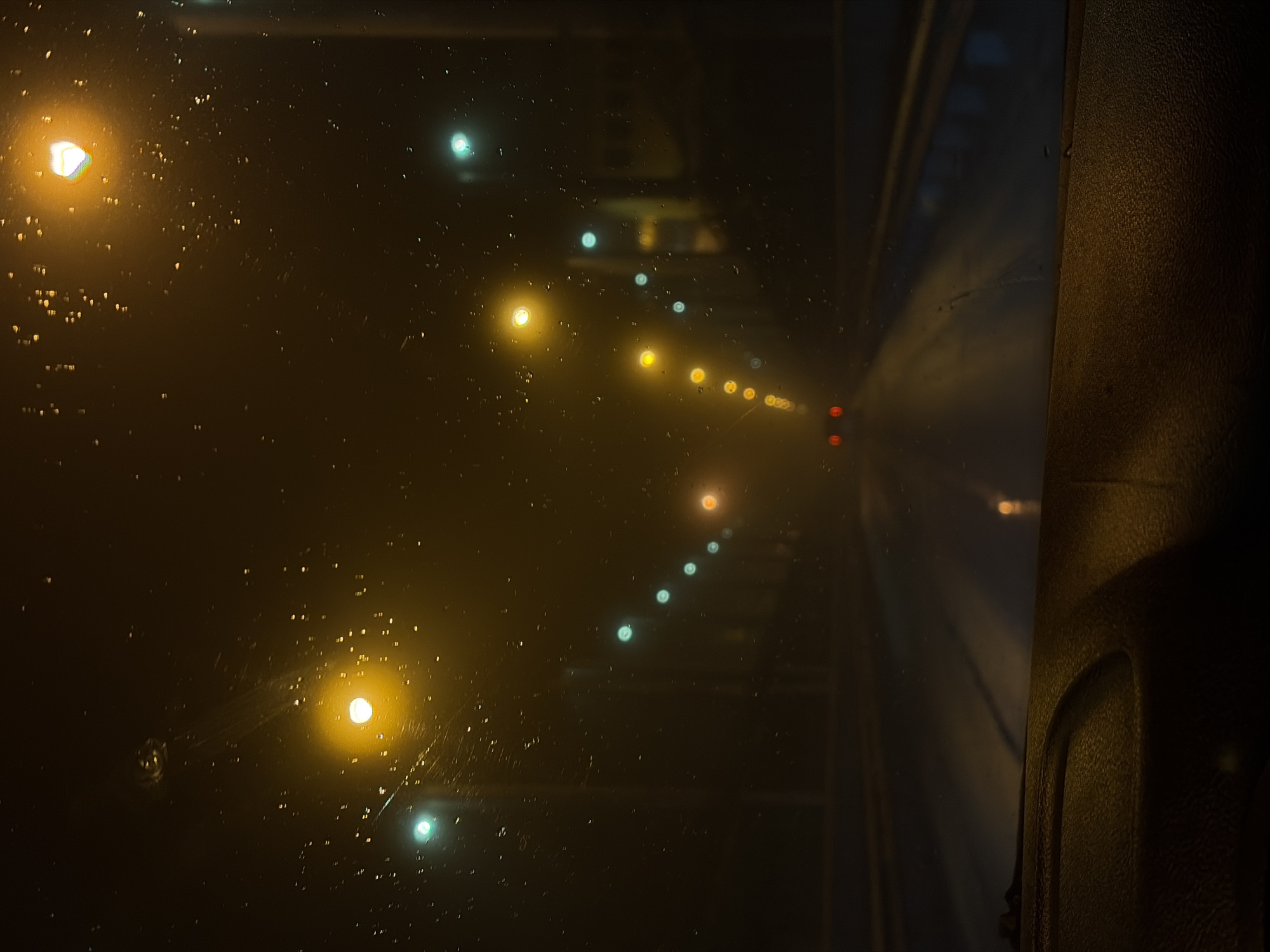 The image depicts a view through a rain-streaked window at night, possibly from inside a vehicle moving through a tunnel in Chaikovsky, Russia. The dominant feature is the presence of numerous droplets on the glass, distorting the light from streetlights and other light sources. The streetlights are visible as blurry, colorful orbs in a line, receding into the distance. There is a dark, textured surface visible on the right side of the frame, likely the interior panel of the vehicle. The overall impression is of motion and nighttime conditions within an urban environment.