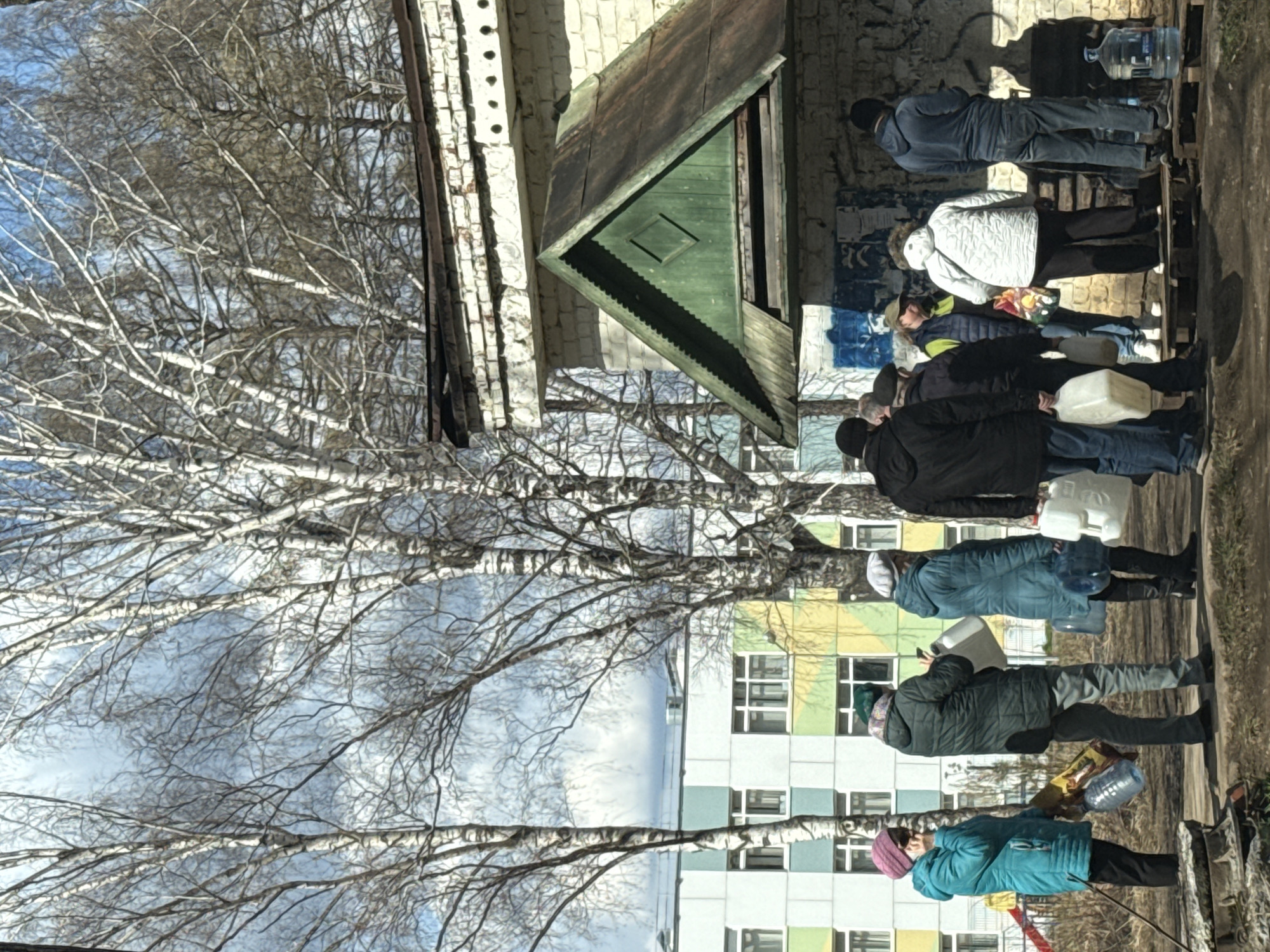A line of people wait, each carrying containers, possibly for water, outside a building in Chaikovsky, Russia. The individuals wear varied clothing, indicating a range of ages and preferences. They are standing on a paved surface next to a building and a row of bare trees, suggesting a cold or early spring season. The architectural style of the building and the overall setting point to an urban area within Chaikovsky. The people appear to be patiently waiting, each holding their container. They interact within the queue, maintaining their place in line. The weather seems clear with visible sky through the tree branches.