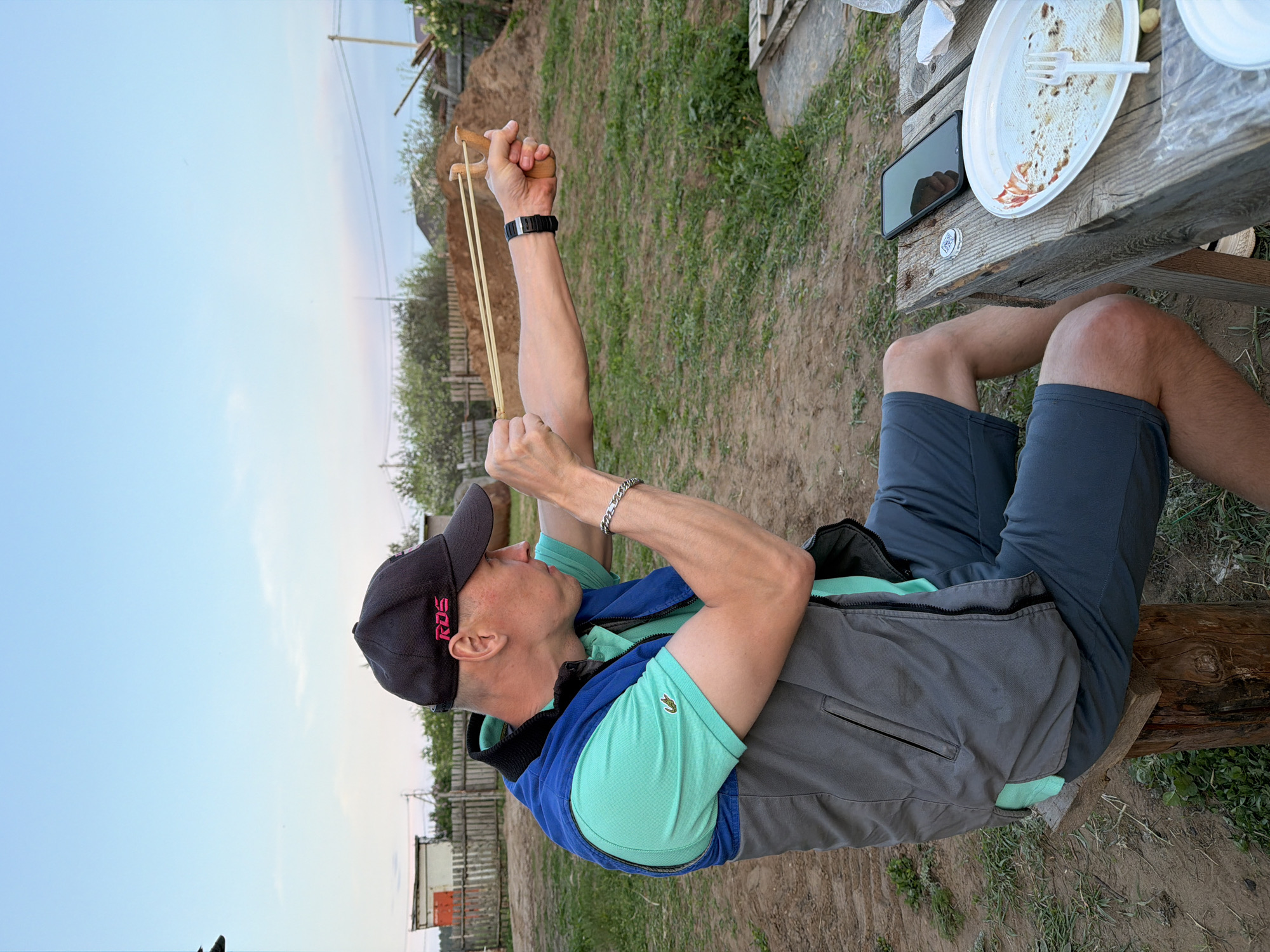 A man sits on a log in an outdoor setting, seemingly at leisure. He is wearing a black baseball cap, a light green polo shirt, a grey vest with blue accents, and dark blue shorts. His left arm is outstretched, pulling back on a wooden slingshot. A black wristwatch is visible on his left wrist. His right hand also grips the slingshot.
Next to him is a weathered wooden table. An empty white plate and a white plastic fork rest on the table, along with a black smartphone and other unidentifiable items. The ground around him appears to be a mix of dirt and grass.
In the background are glimpses of trees, structures, and power lines. Based on the information provided, this location could be in or near the city of Chaikovsky, Russia.