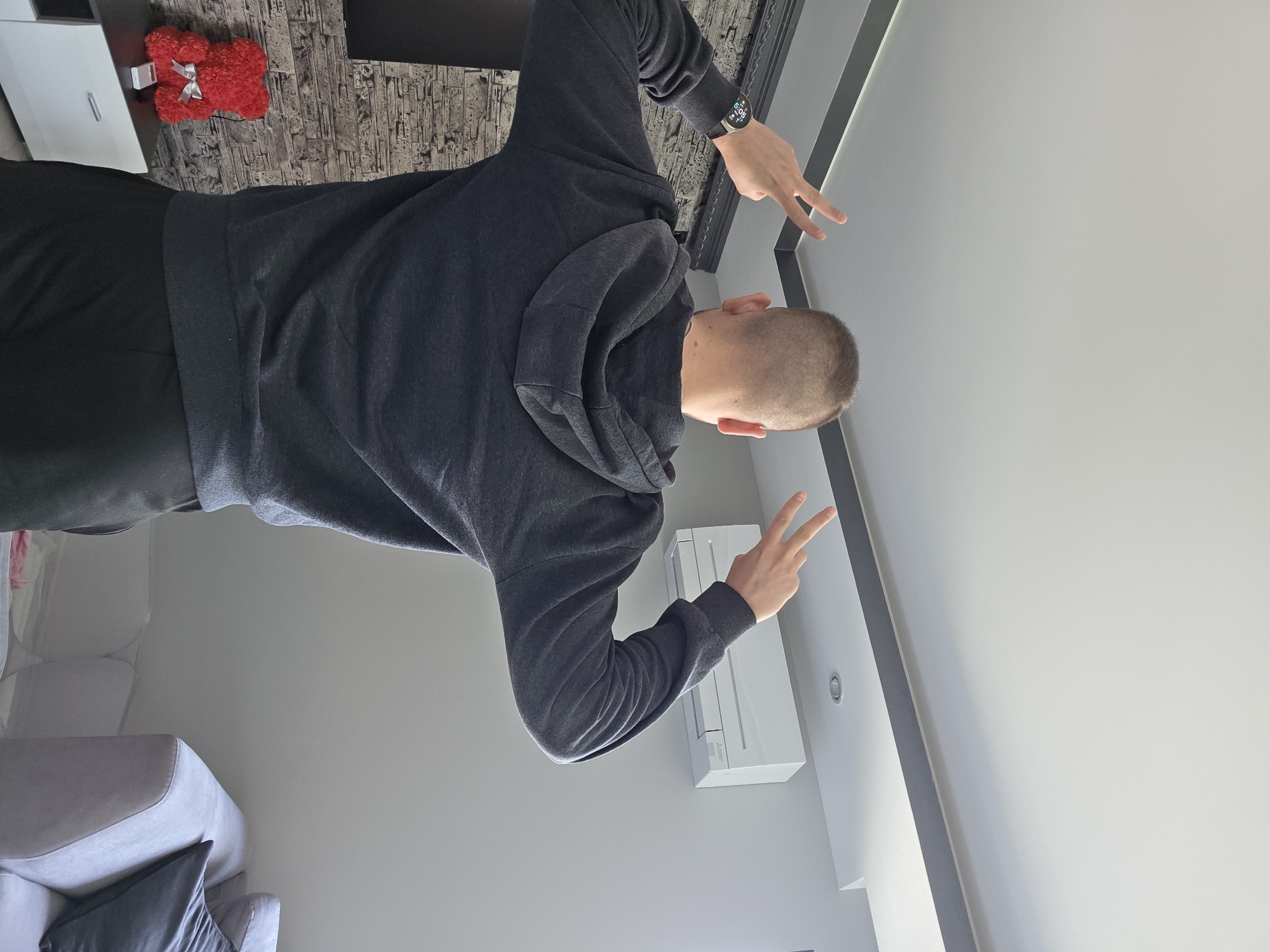A man with a shaved head, wearing a dark gray hooded sweatshirt and dark pants, is making the peace sign with both hands. He is positioned indoors against a light gray wall with an air conditioner mounted on it. Behind him, there is a dark gray-patterned surface, possibly a wall or furniture, and a small red floral arrangement on top of a white cabinet or shelf. A gray sofa with pillows is visible in the lower left corner. The location appears to be inside a modern residential building, but there is no verifiable connection to Yagodovo, Bulgaria.