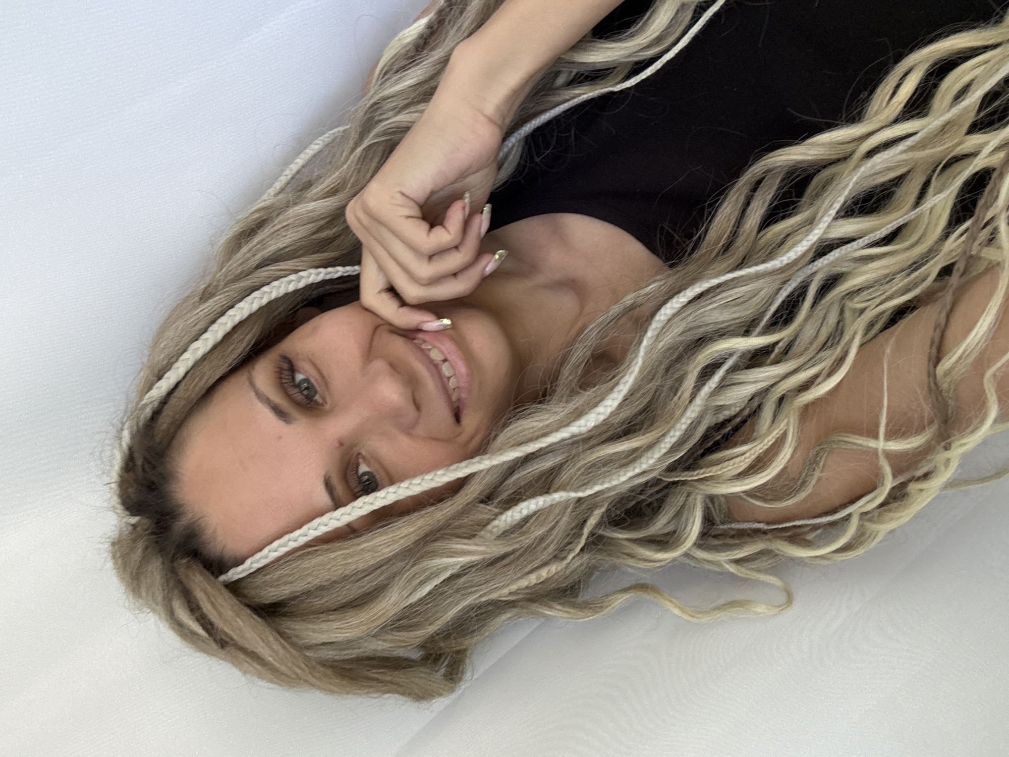 A woman with blonde braided hair, wearing a black top, is seen from a top-down perspective against a white background. She is smiling slightly, with her hand near her face, showing painted nails. The image appears to have been taken indoors. The location is not discernable from the image.